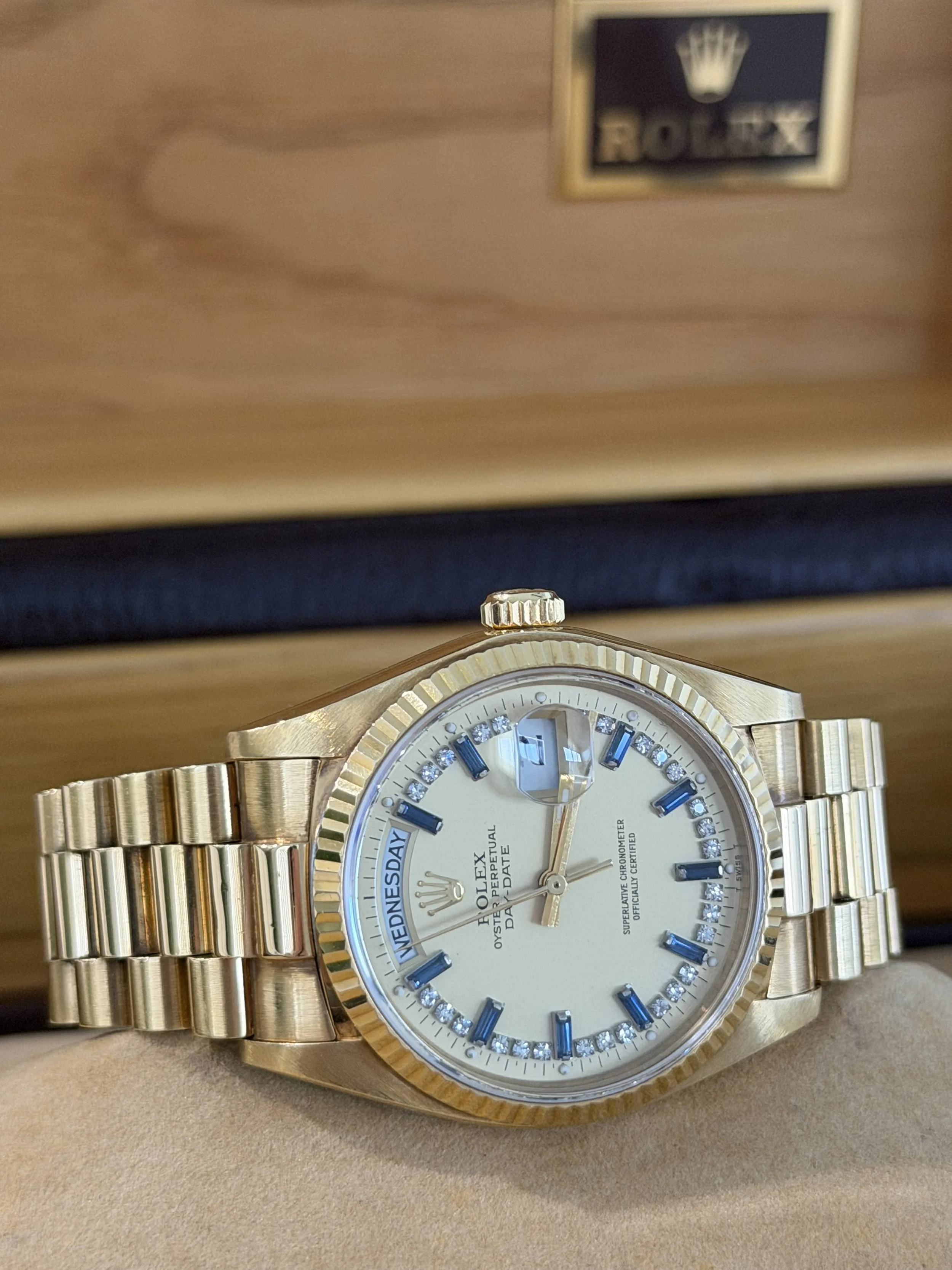 A gold Rolex wristwatch with a white face, blue Roman numeral hour markers, and diamond accents, placed on a beige surface.