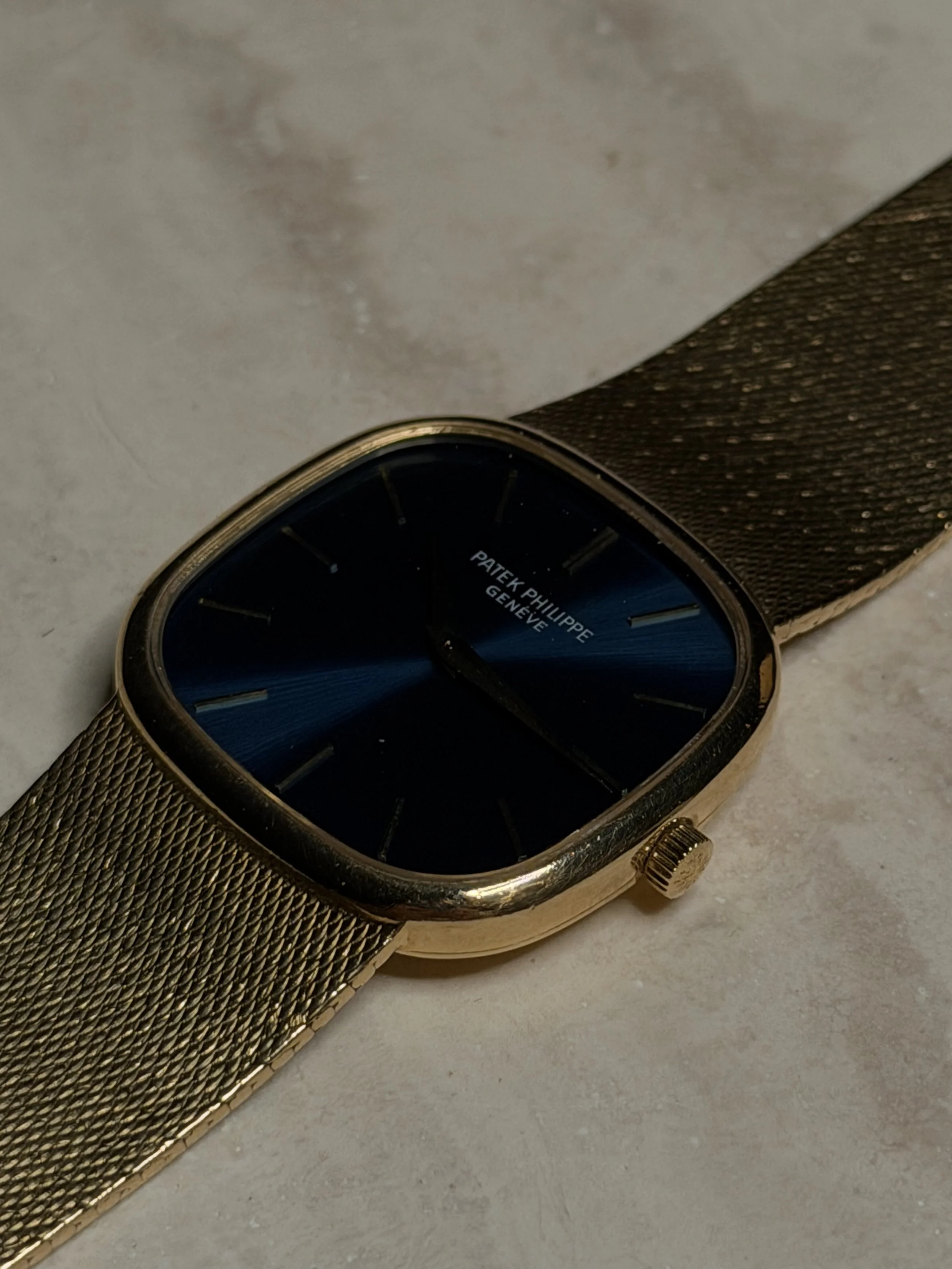 A gold-tone wristwatch with a black face, gold hour markers, and the words 'Patek Philippe Genève' on the dial, resting on a textured gold bracelet and a light-colored surface.