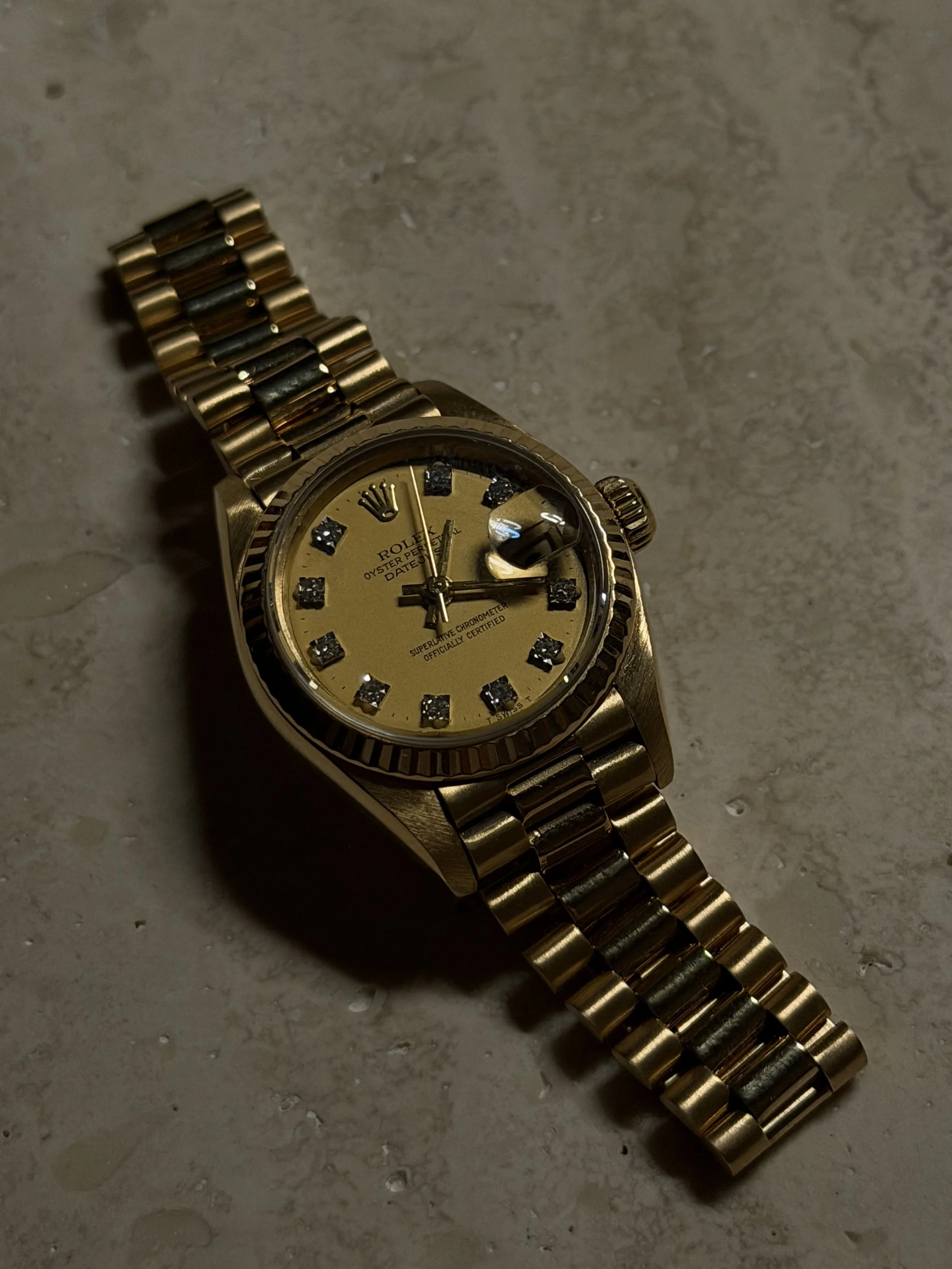 A gold Rolex wristwatch with a gold band resting on a beige textured surface.