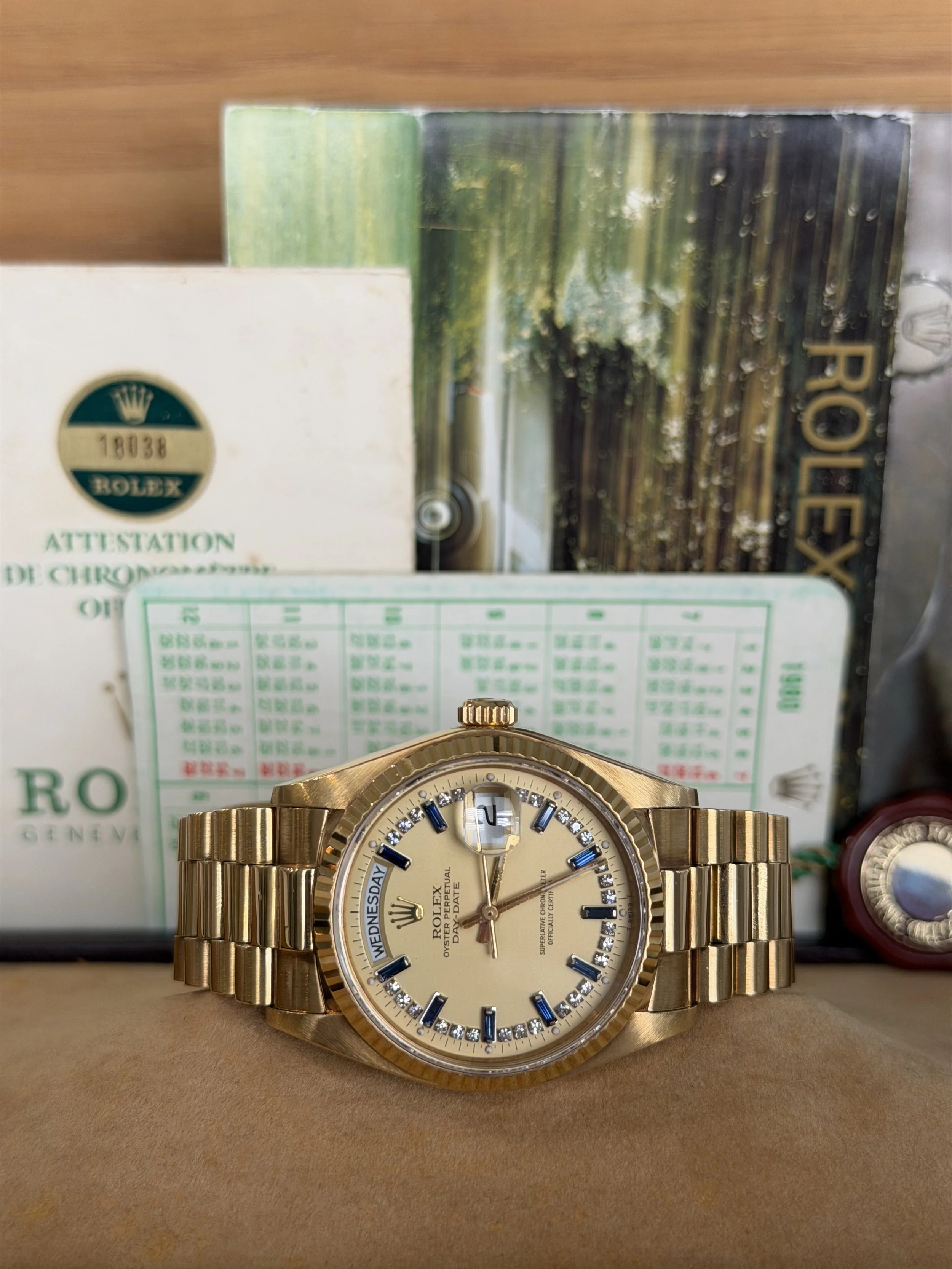 A gold Rolex wristwatch with a round face, blue hour markers, and diamond accents, placed in front of a display box and certification papers.