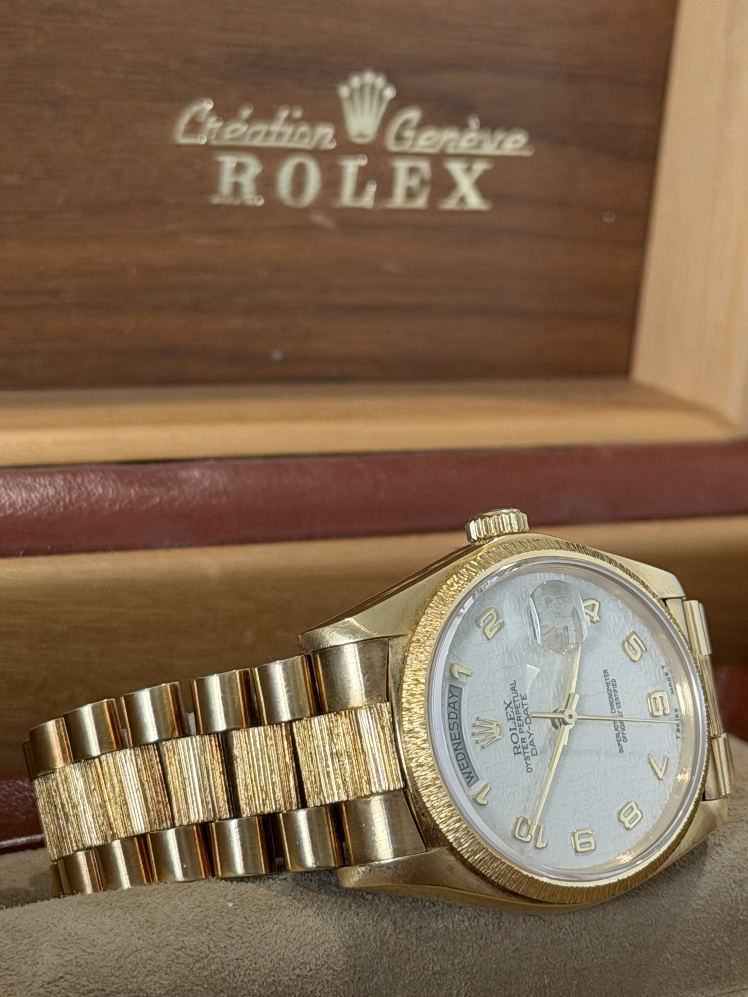 A gold Rolex wristwatch with a metal band and a white face displaying the days of the week, sitting in front of a wooden box with the Rolex logo and the words "Oyster Perpetual Day-Date".