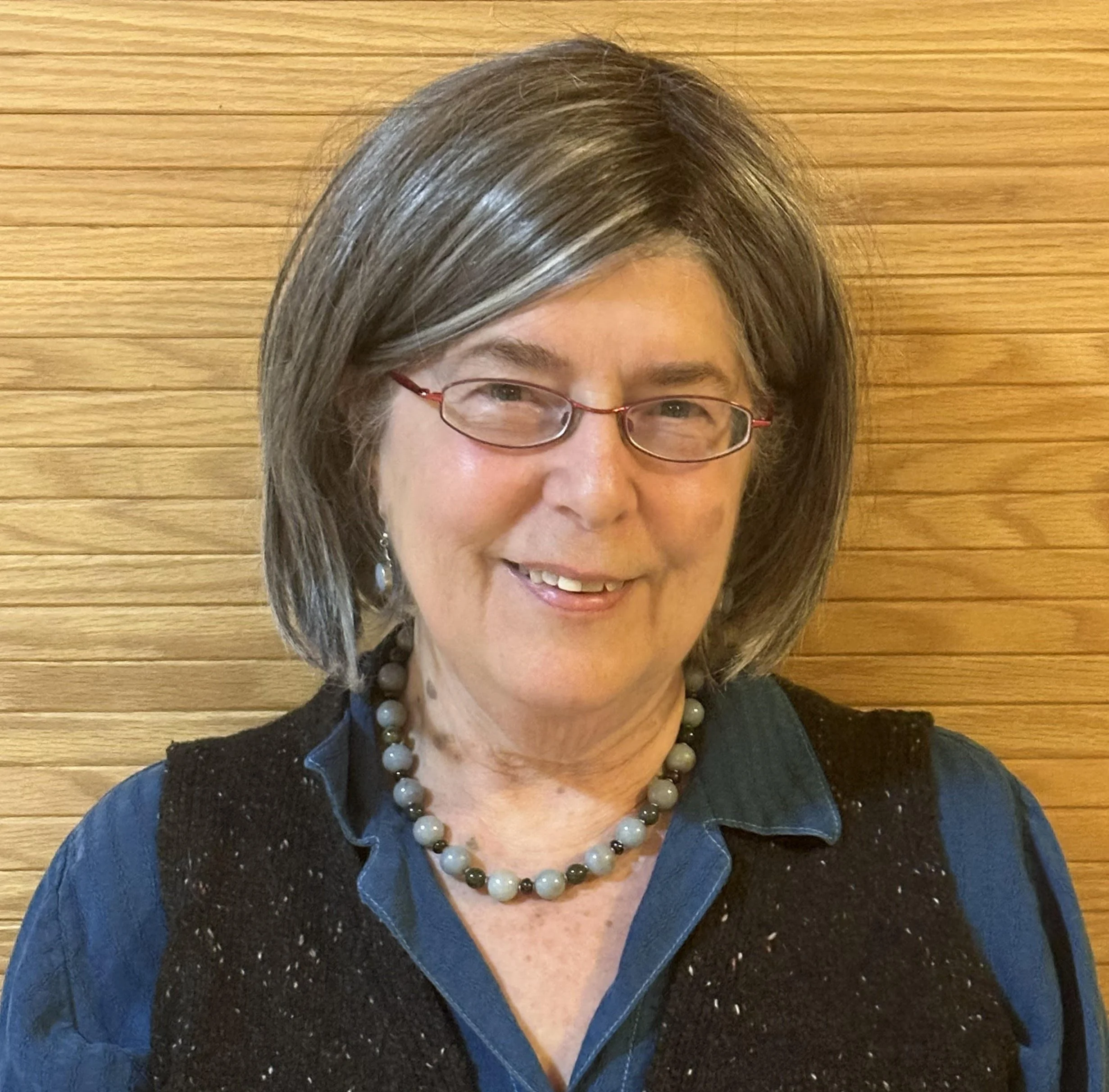 Close-up of a smiling person wearing glasses, a dark blue top, and a multi-colored bead necklace, standing in front of a wooden wall.