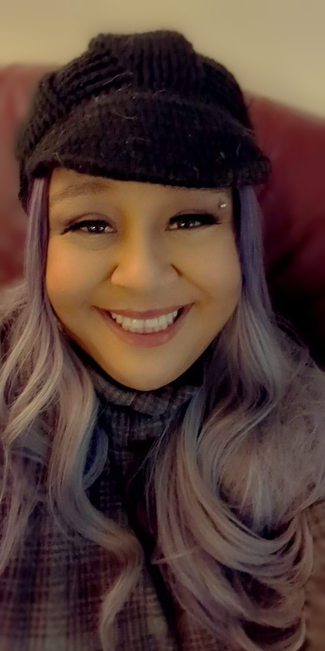 A Black person with long, wavy, light purple hair wearing a black knit beanie and a plaid shirt, smiling at the camera.