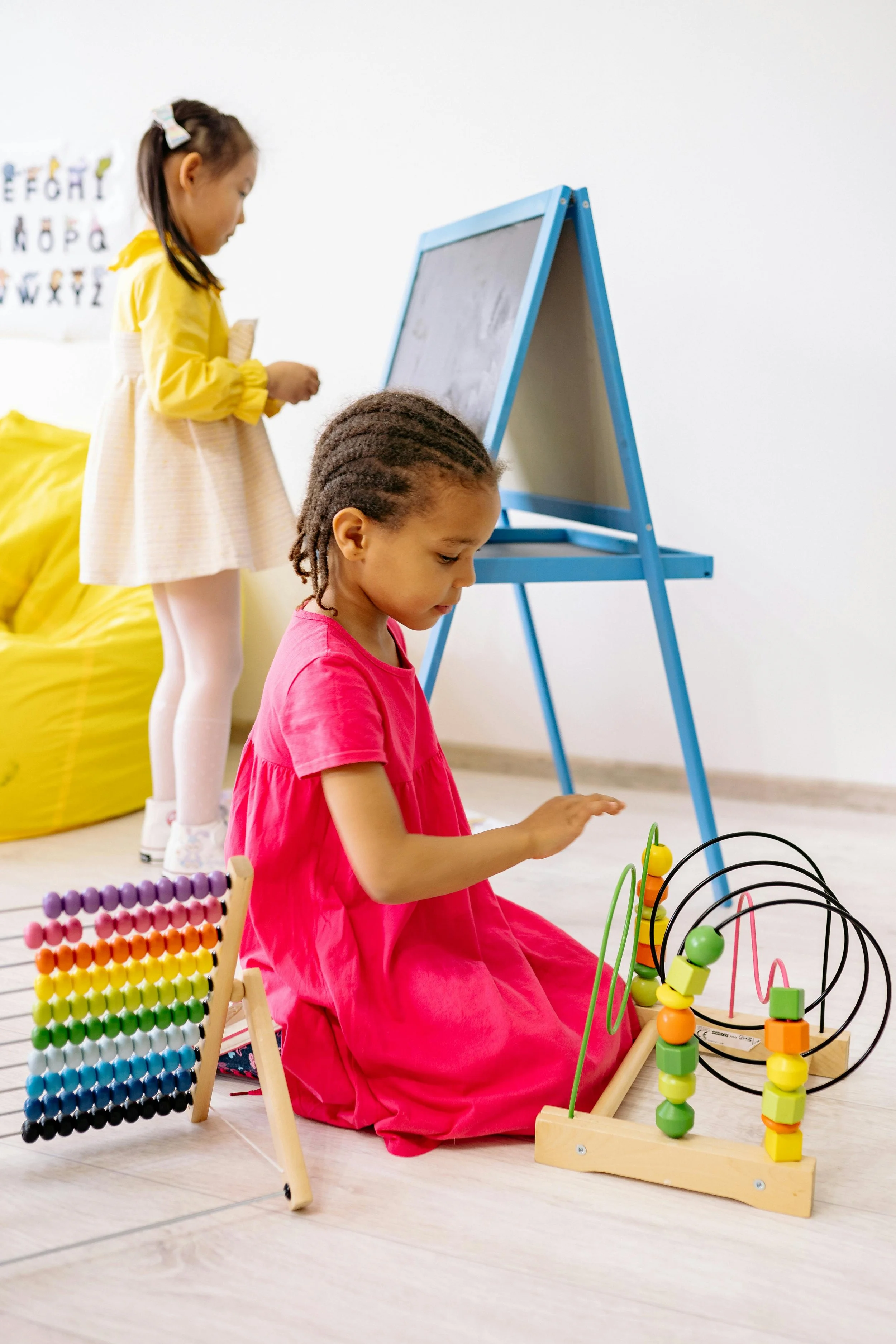Two young girls playing with educational toys on a light wooden floor, one sitting and the other standing, in a bright room with a white wall, a blue easel, a colorful bead maze, and an alphabet poster in the background.