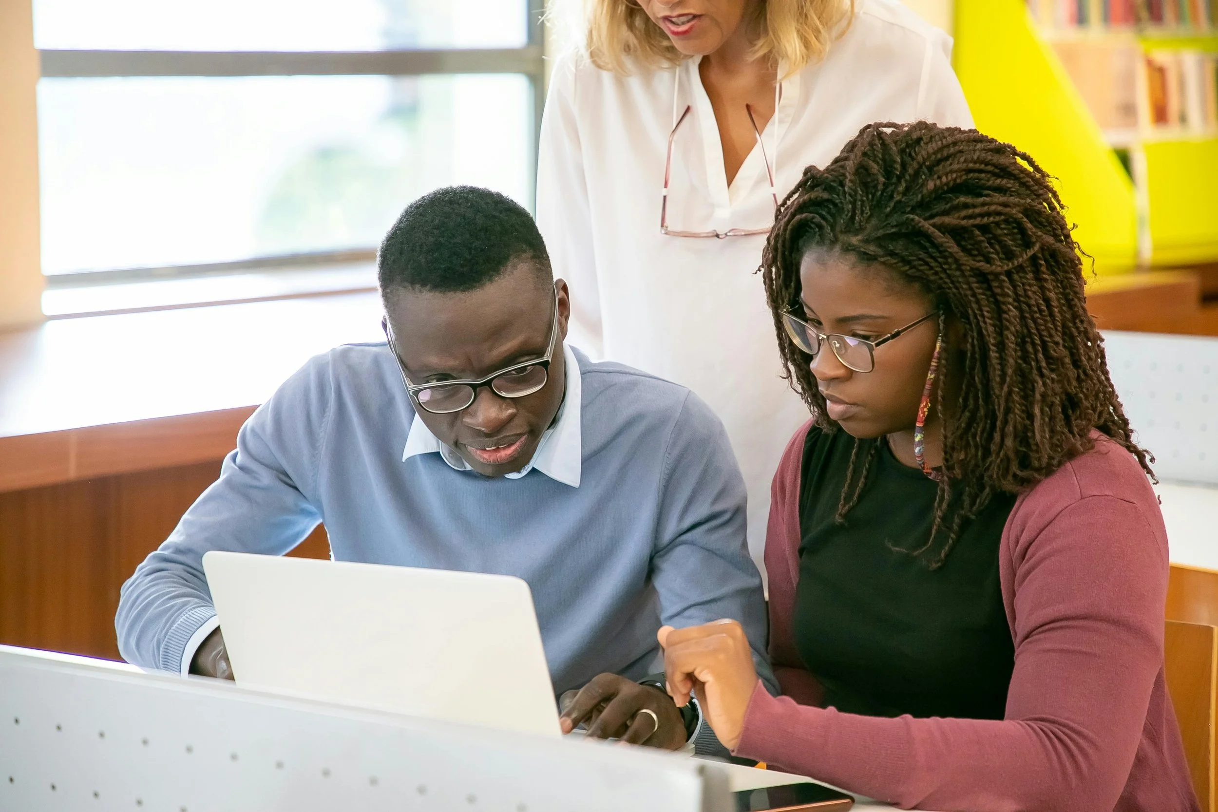 Two Black people, one with short curly hair and glasses and the other with long braids, are sitting together at a desk, looking at a laptop screen. An older person with blonde hair is standing behind them, leaning over to look at the laptop.