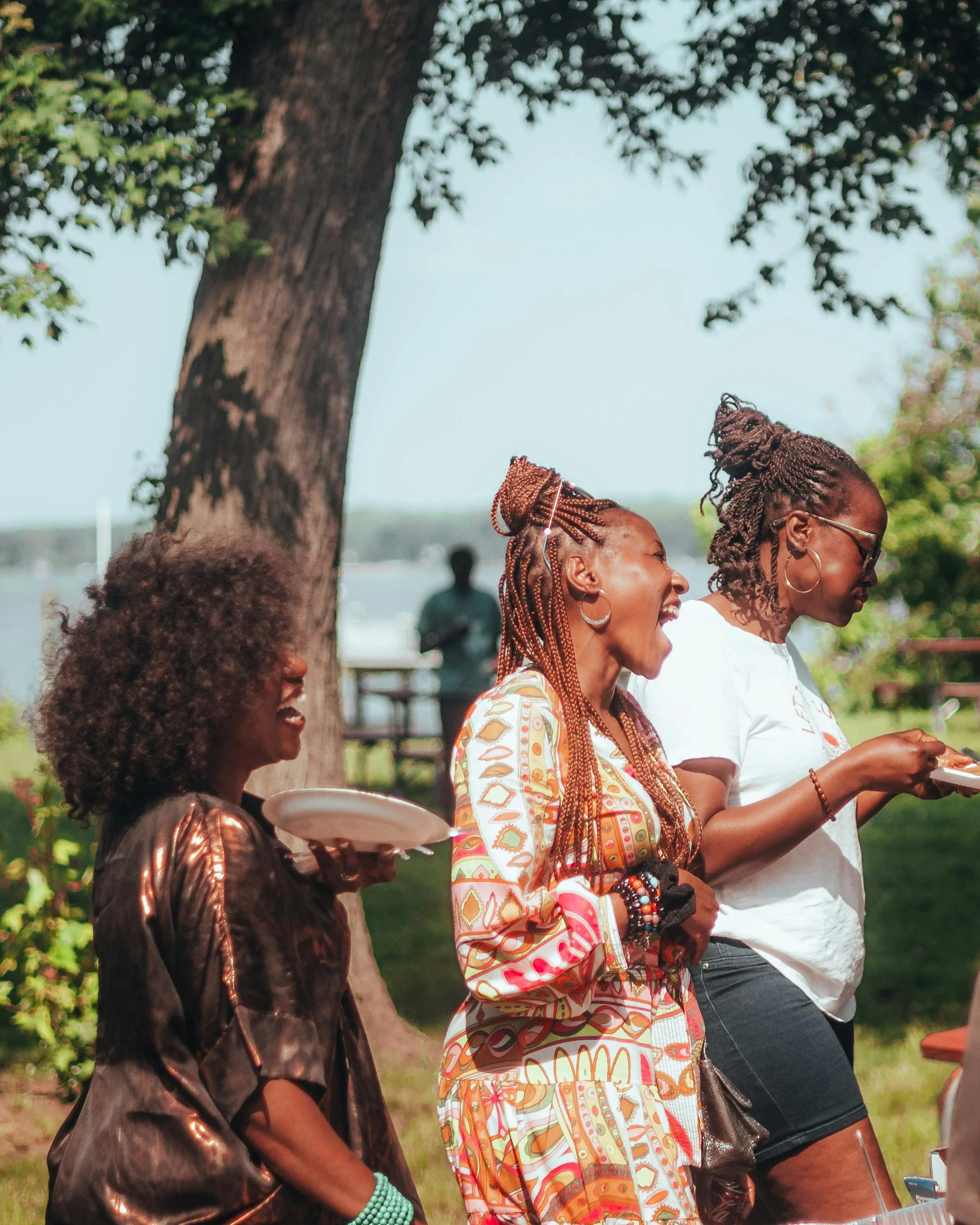 A group of Black people standing outdoors near a large tree, enjoying a social gathering, with some holding plates of food and others smiling and engaged in conversation.