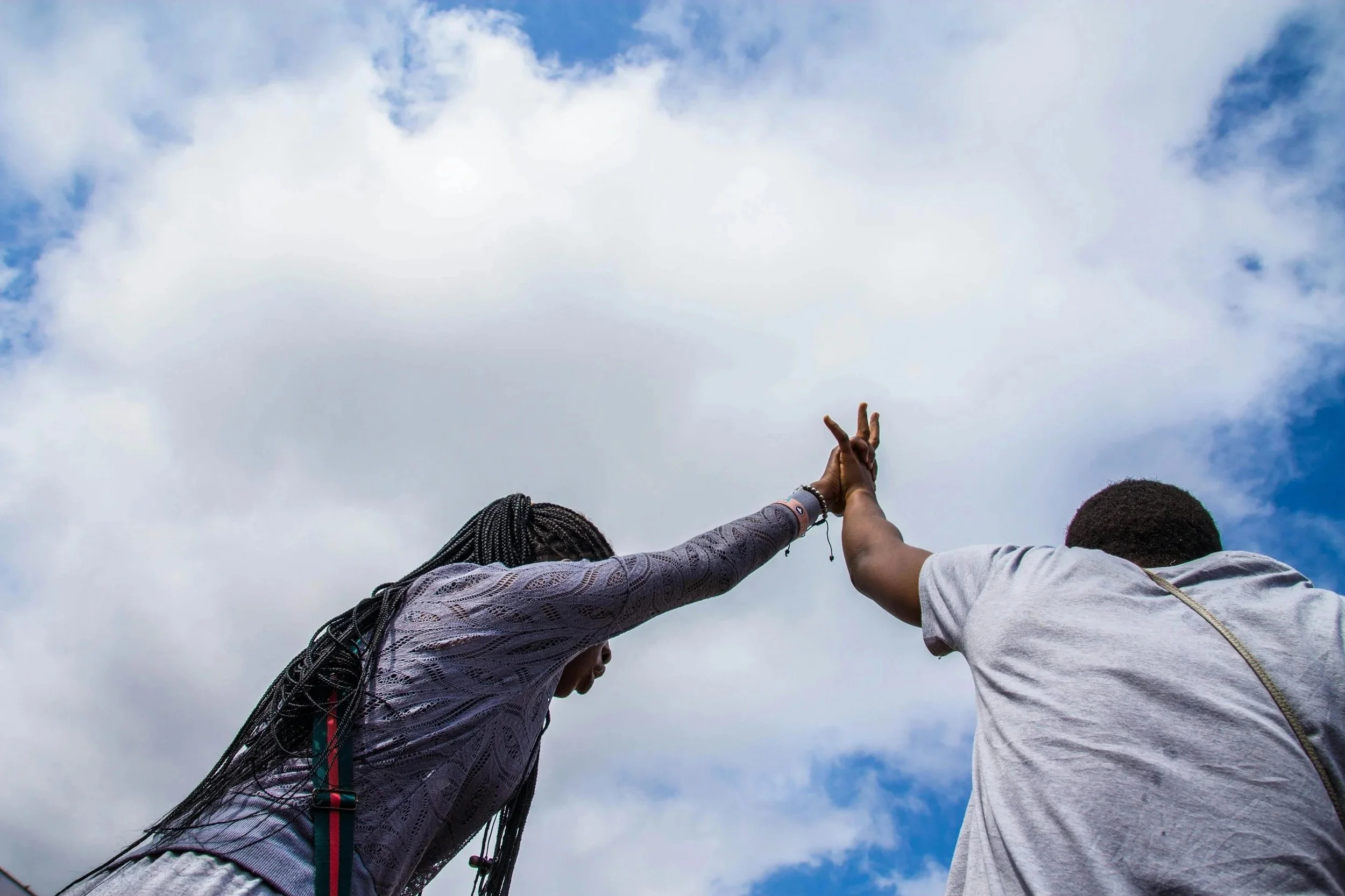 Two people reaching upward with their hands, touching or overlapping, against a cloudy sky in the background.