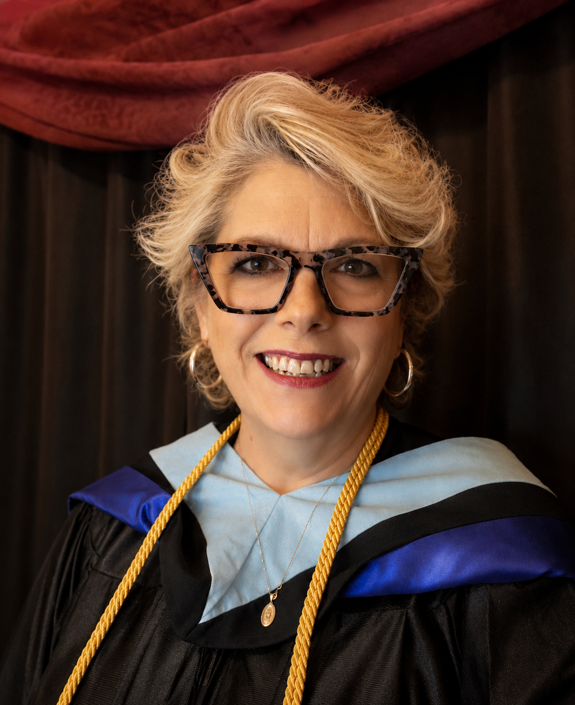 A person in graduation attire with a black gown, light blue hood, and gold stole, wearing large glasses and smiling at the camera wearing hoop earrings, a necklace, and a gold honor cord.