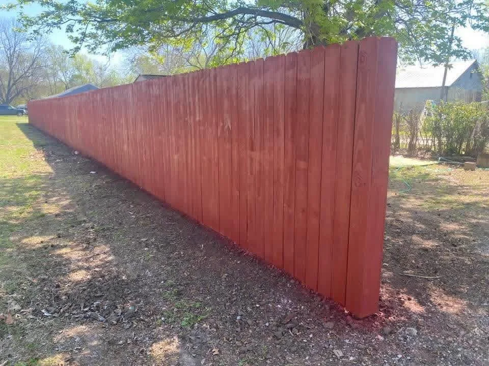 A freshly painted red wooden fence running along a yard with trees, shrubs, and a house in the background.