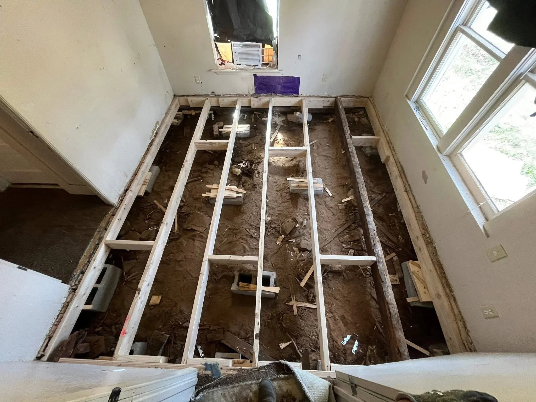 Room under renovation with exposed wooden floor framing, a window on the right, and construction debris on the dirt floor.