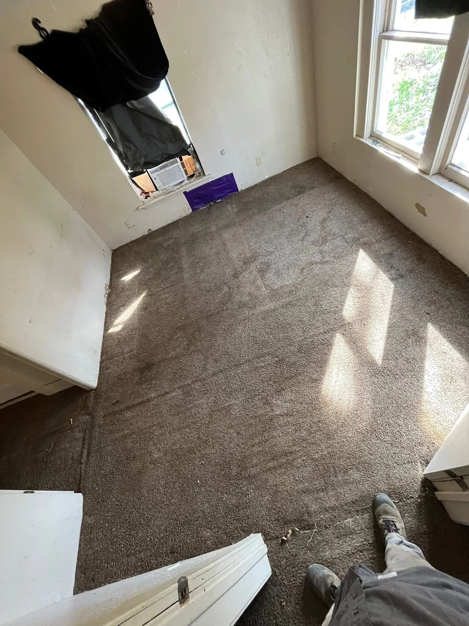 A small, empty room with beige carpet, white walls, a window with black curtains, and a person standing at the bottom right corner wearing gray pants and sneakers. There are some objects like a purple box, an air conditioning unit, and some loose items on the window sill and floor.