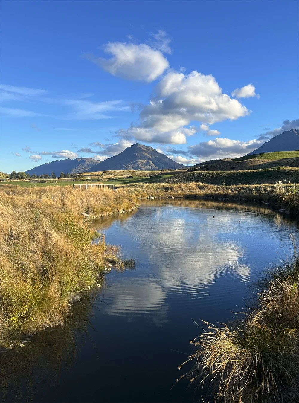 A scenic landscape featuring a calm river reflecting the sky with white clouds, surrounded by yellowish grass and rolling green hills, with mountain peaks in the background under a blue sky.