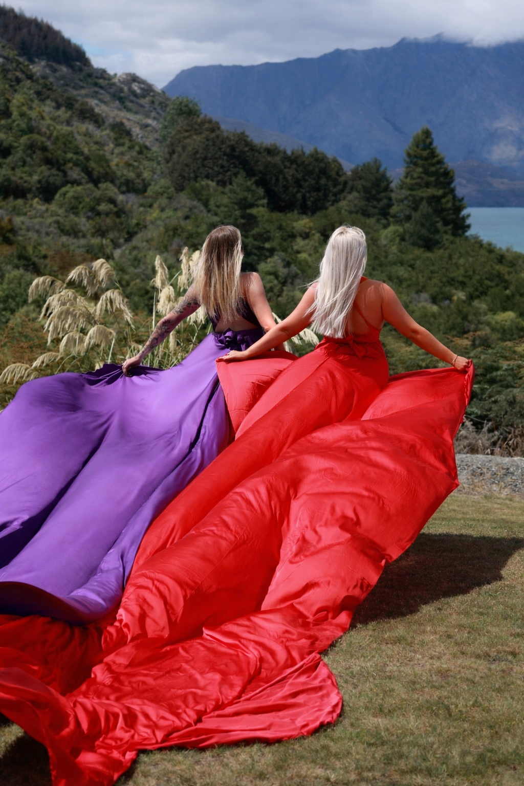 Two women in colorful dresses, one purple and one red, walking outdoors on a grassy area with a scenic mountain and lake background.