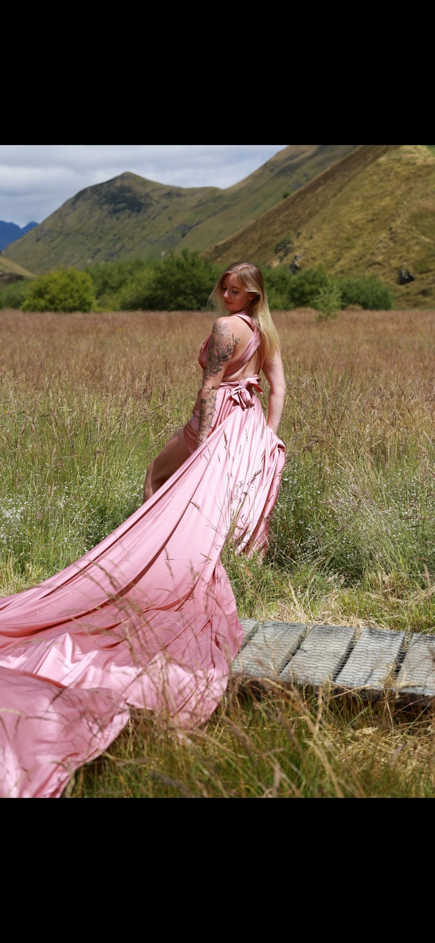 Woman in pink gown walking through grassy field with hills and mountains in the background.