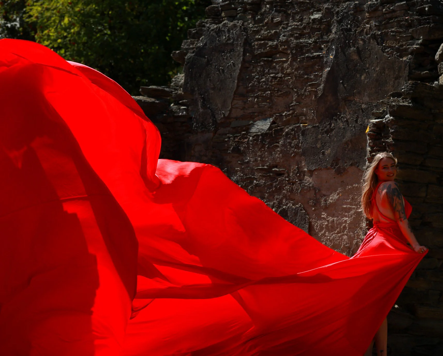 A woman with long blonde hair and tattoos on her arm, wearing a red dress, sitting against a stone wall with a red fabric flowing around her.