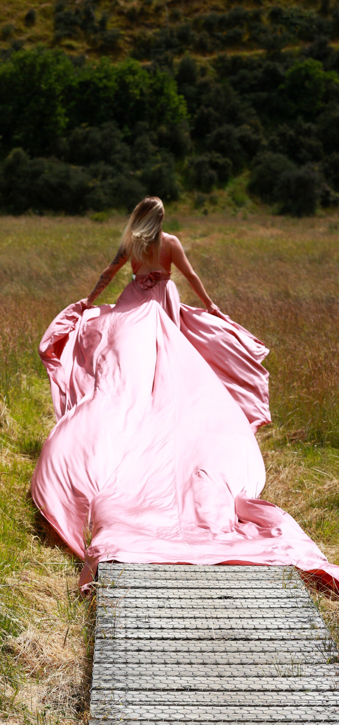 Woman in a flowing pink gown standing on a wooden bridge in a grassy field with trees and hills in the background.