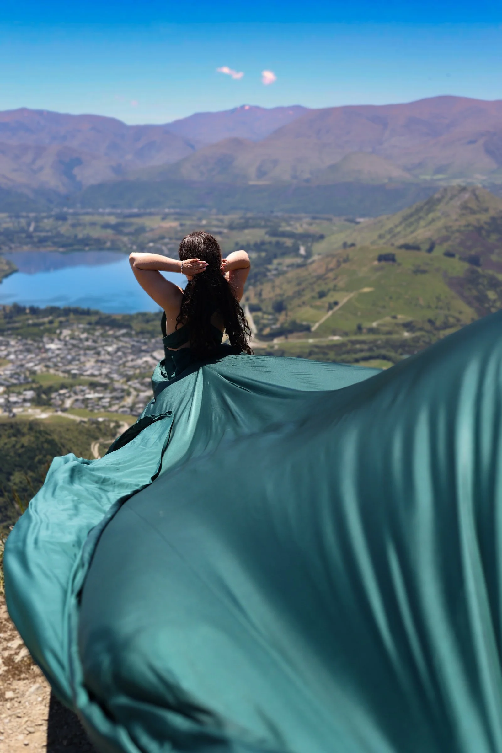 A woman with long dark hair sitting on a grassy hilltop overlooking a valley with mountains, a lake, and a small town, with a blue sky and a few clouds in the background.