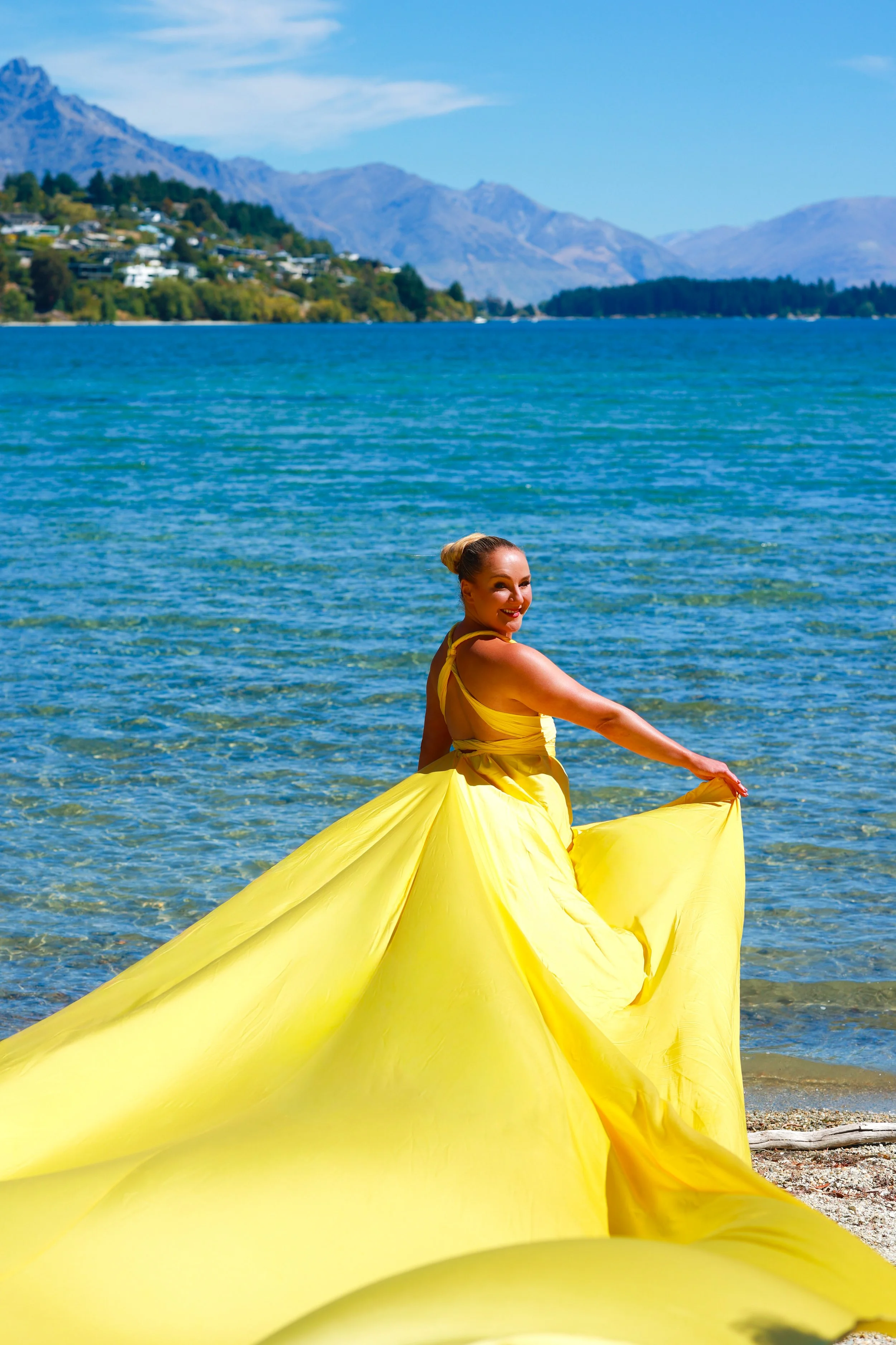 A woman in a yellow dress standing by the water, with mountains and houses in the background.