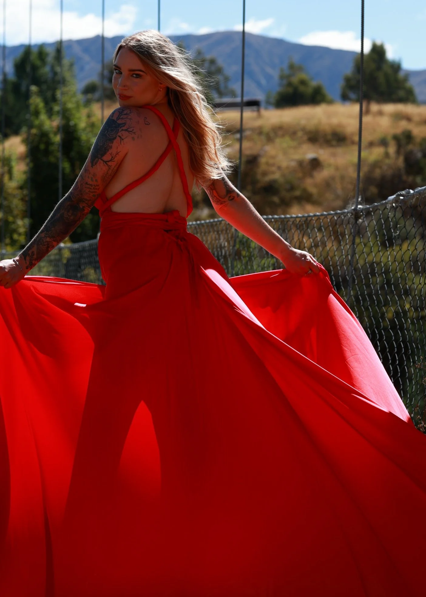 A woman with tattoos on her left arm and shoulder, wearing a red dress with a flowing skirt, outdoors with mountains and trees in the background.