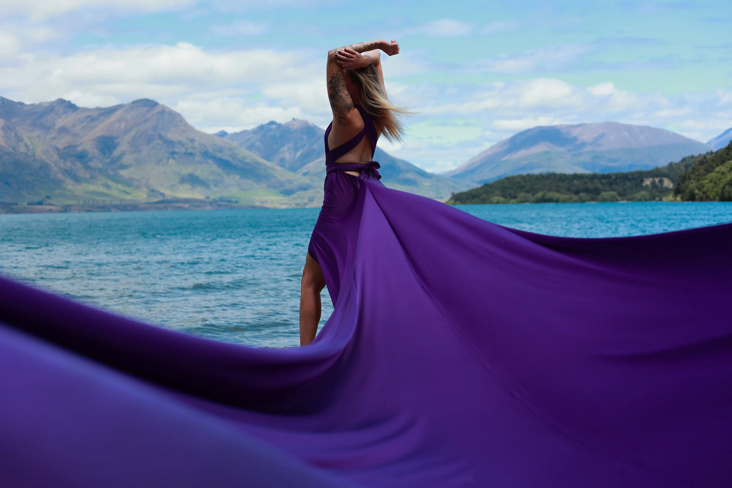 A woman in a purple dress with a flowing train stands at the edge of a lake with mountains in the background, seen through a frame of purple fabric.