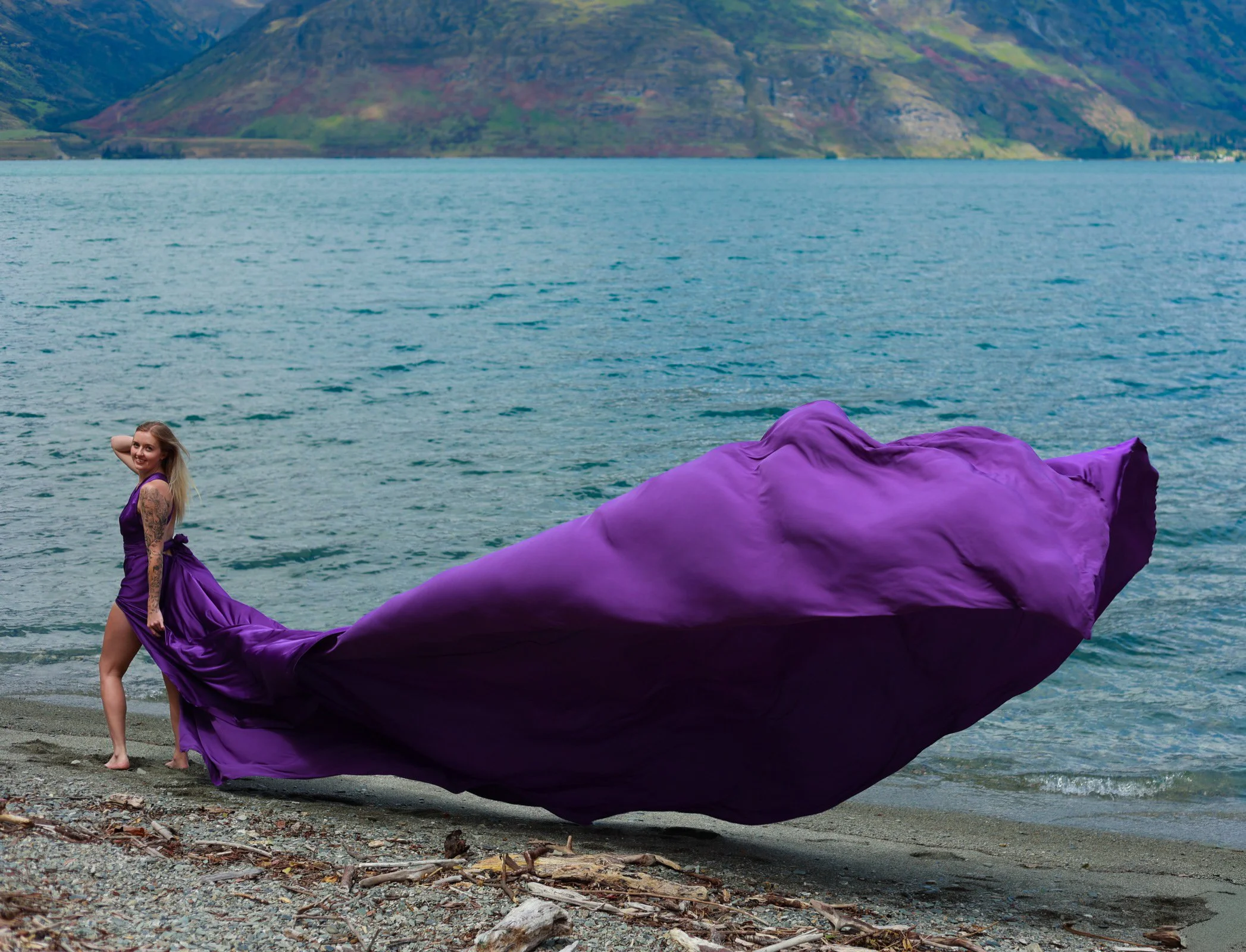 A woman in a purple dress standing on a rocky beach by a lake with mountains in the background. The dress has a long train flowing onto the ground.