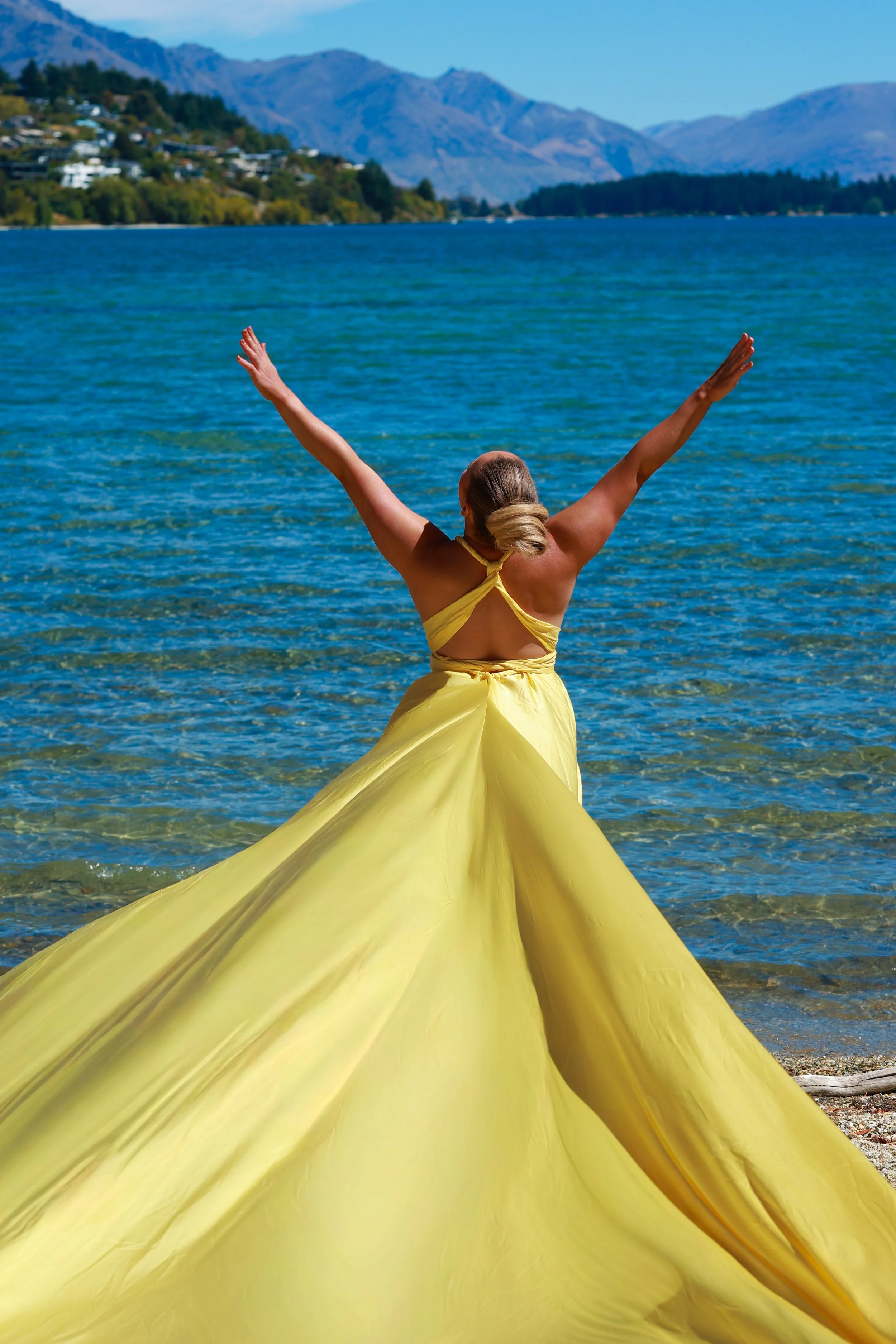 A woman in a yellow dress standing in shallow water with arms raised, facing a lake with mountains and houses in the background during daytime.