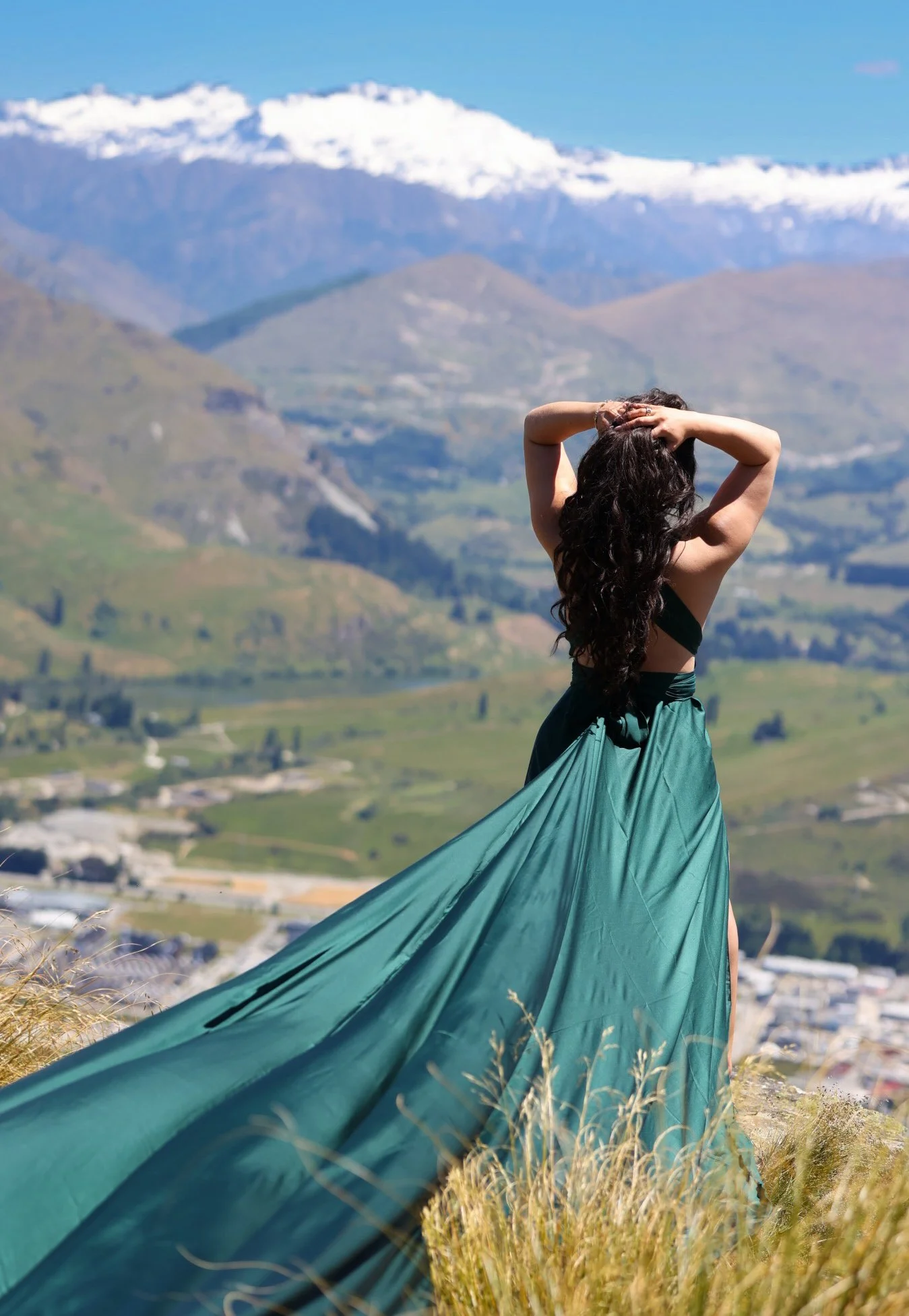 A woman with long dark curly hair standing outdoors on a hillside, wearing a flowing teal dress, with a background of mountains, some with snow caps, and a blue sky.