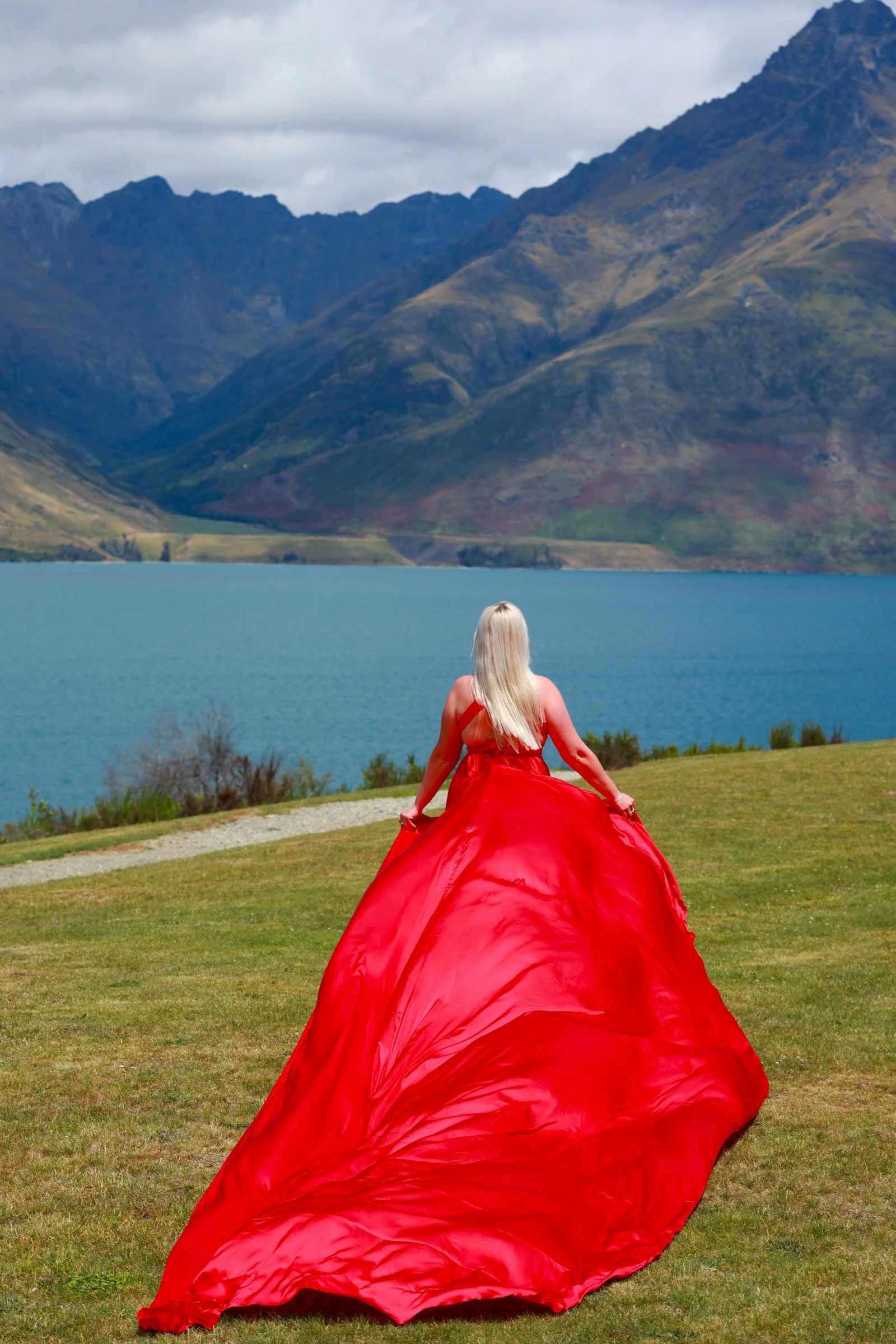 A woman with long blonde hair in a red dress sitting on green grass by a lake, with mountains and cloudy sky in the background.