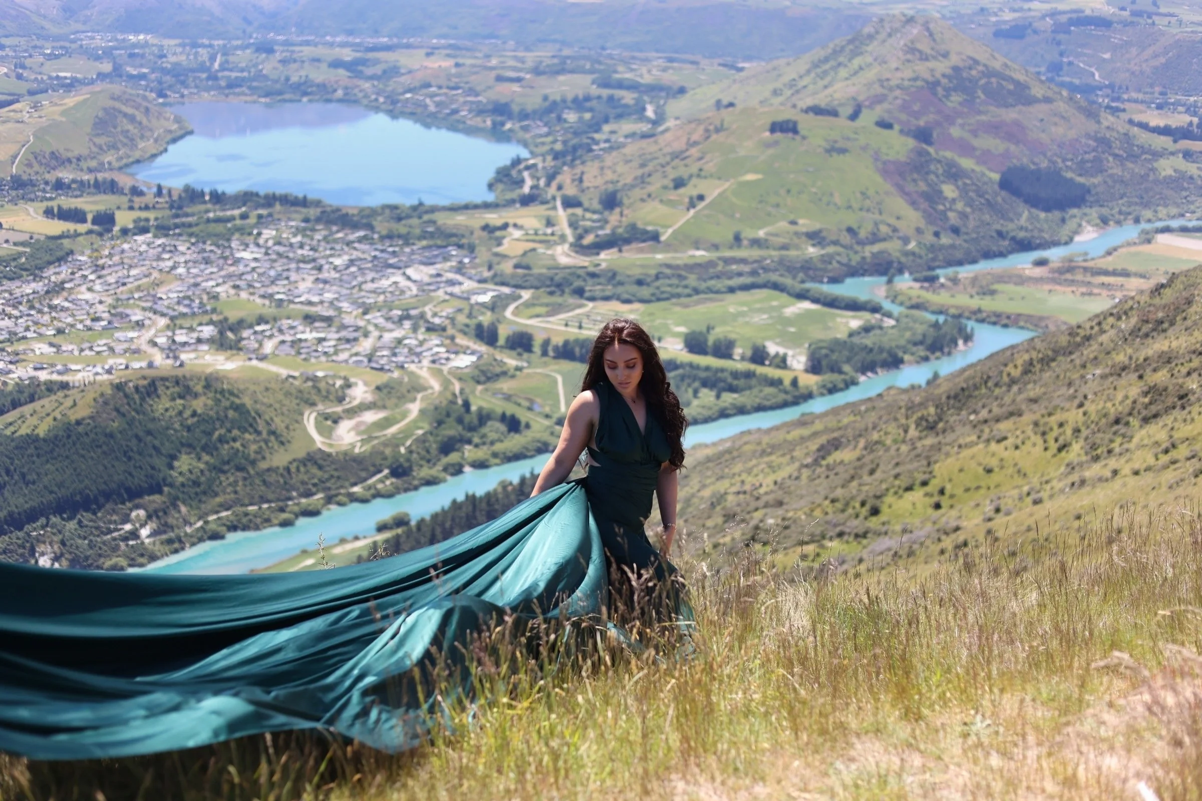 A woman in a dark green dress standing on a hillside with a flowing fabric in her hand, overlooking a scenic landscape of a lake, river, hills, and a small town.