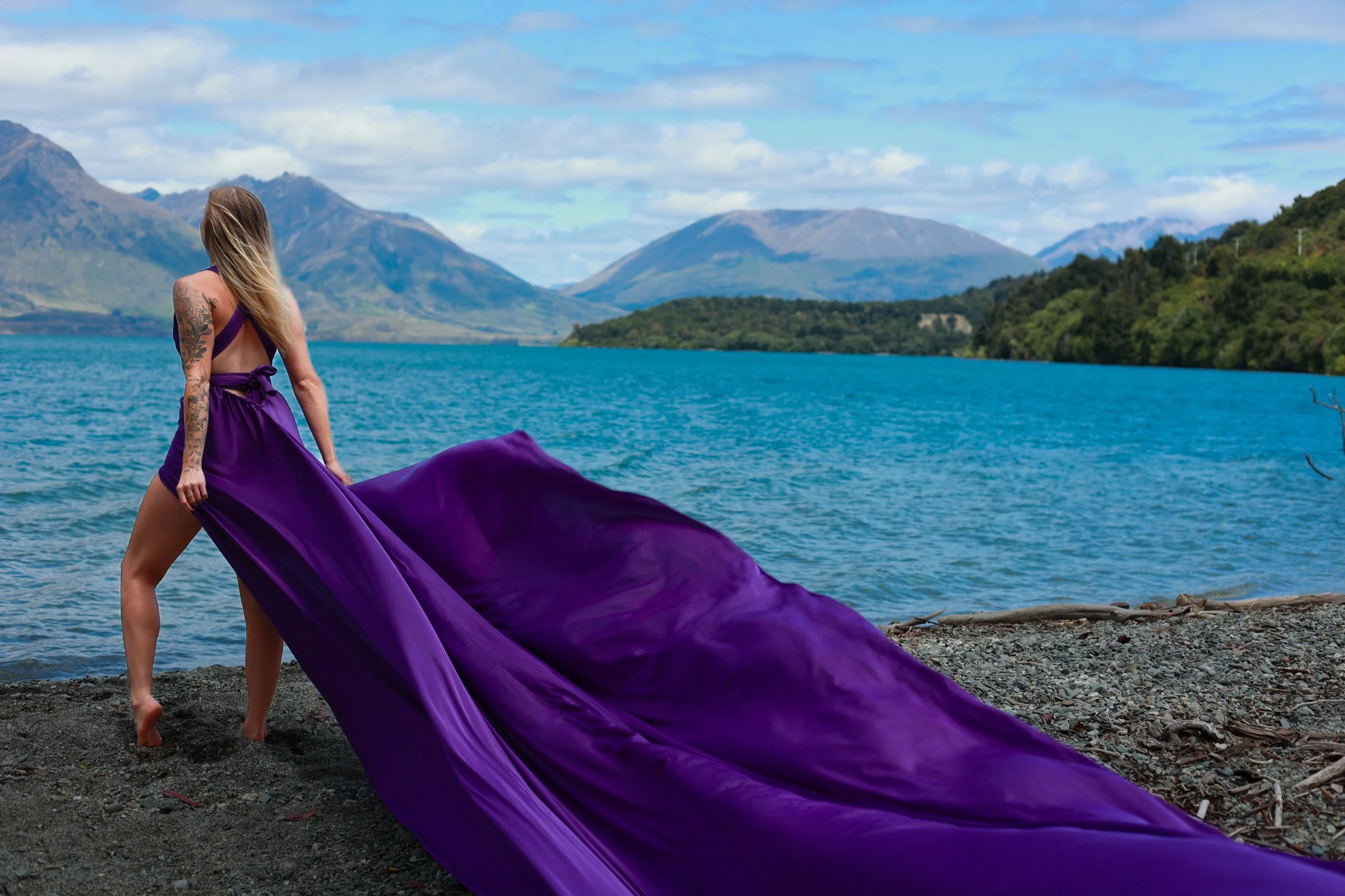 Woman with long hair, wearing a purple dress with open back tattoos, standing barefoot on a rocky shoreline by a lake, with mountains and a partly cloudy sky in the background.