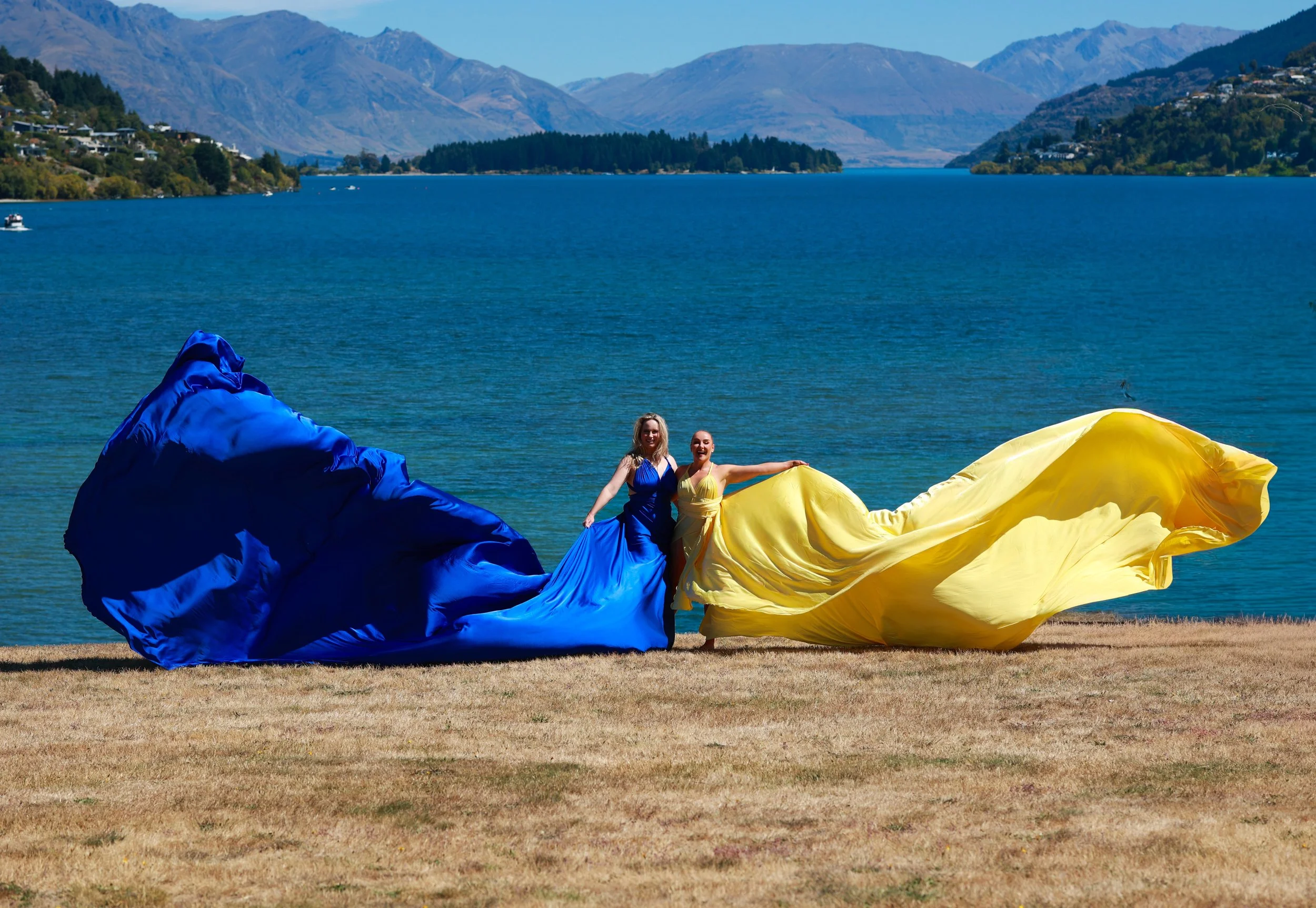 Two women in colorful dresses are standing outdoors near a lake, holding large pieces of blue and yellow fabric that are fluttering in the wind. The background features a mountain range and a partly cloudy sky.