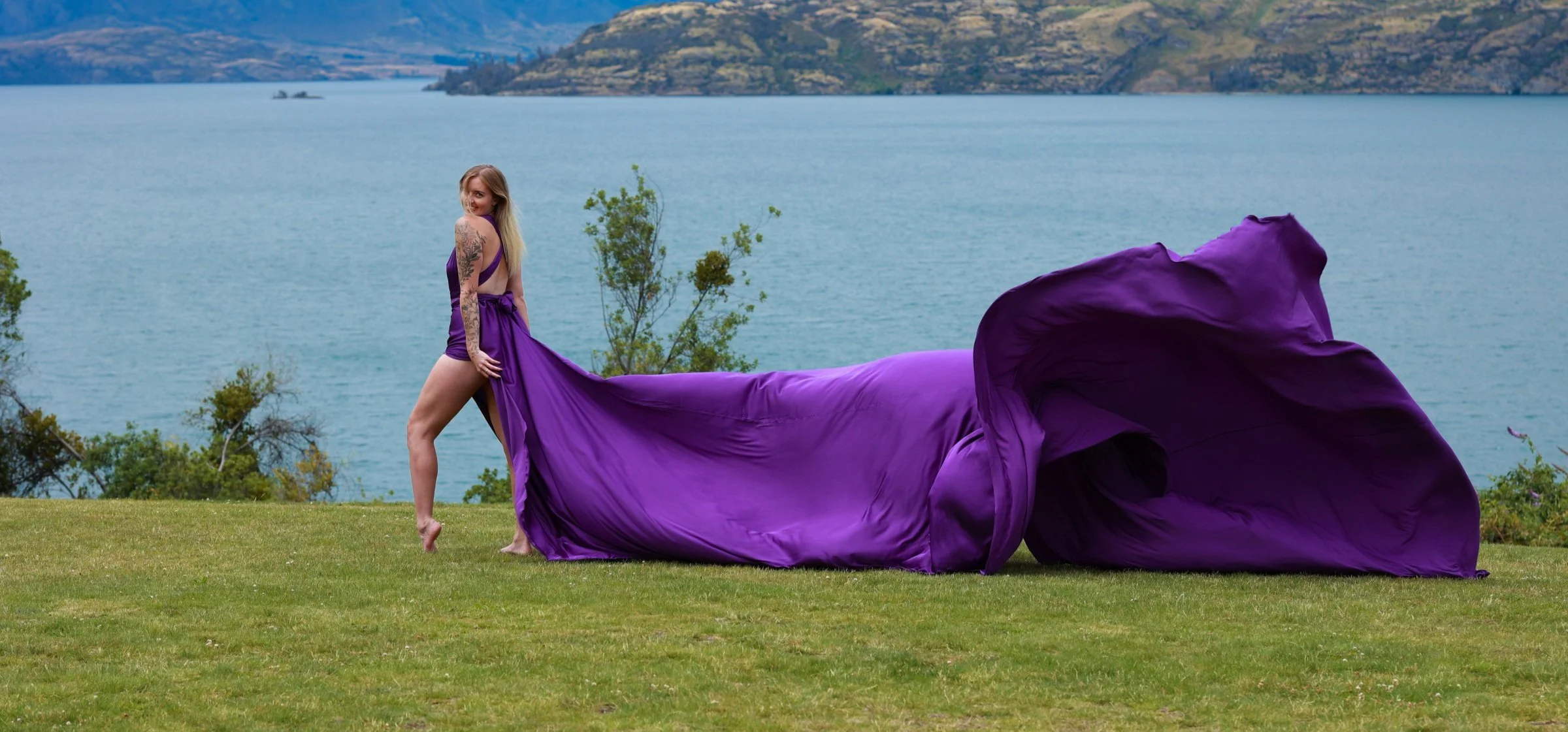 A woman with tattoos wearing a purple gown with a long train, standing on a grassy field near a body of water with hills in the background.