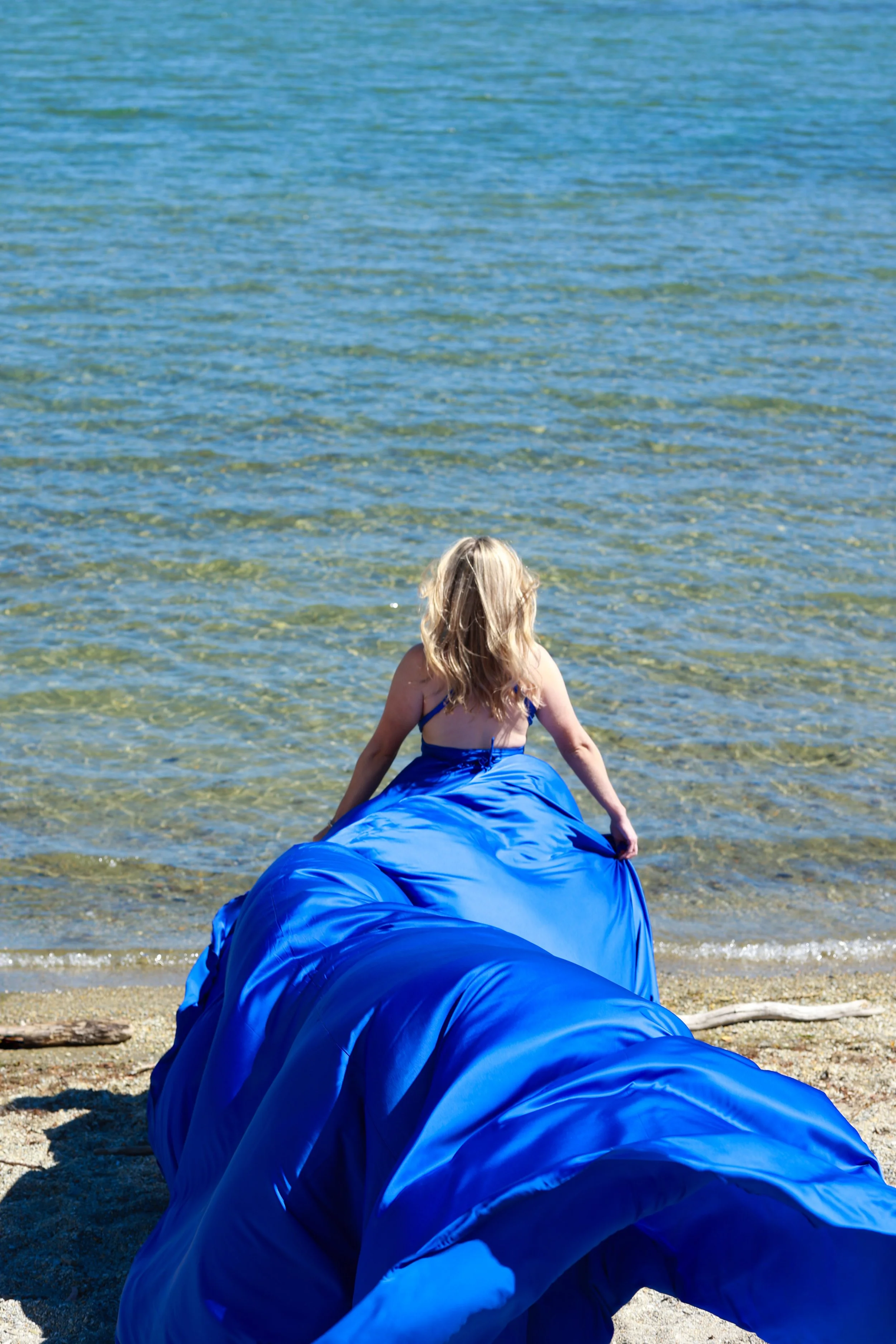 A young girl with blonde hair wearing a blue swimsuit sitting on a blue inflatable raft at the edge of the ocean, facing away from the camera, with water and a sandy beach in the background.