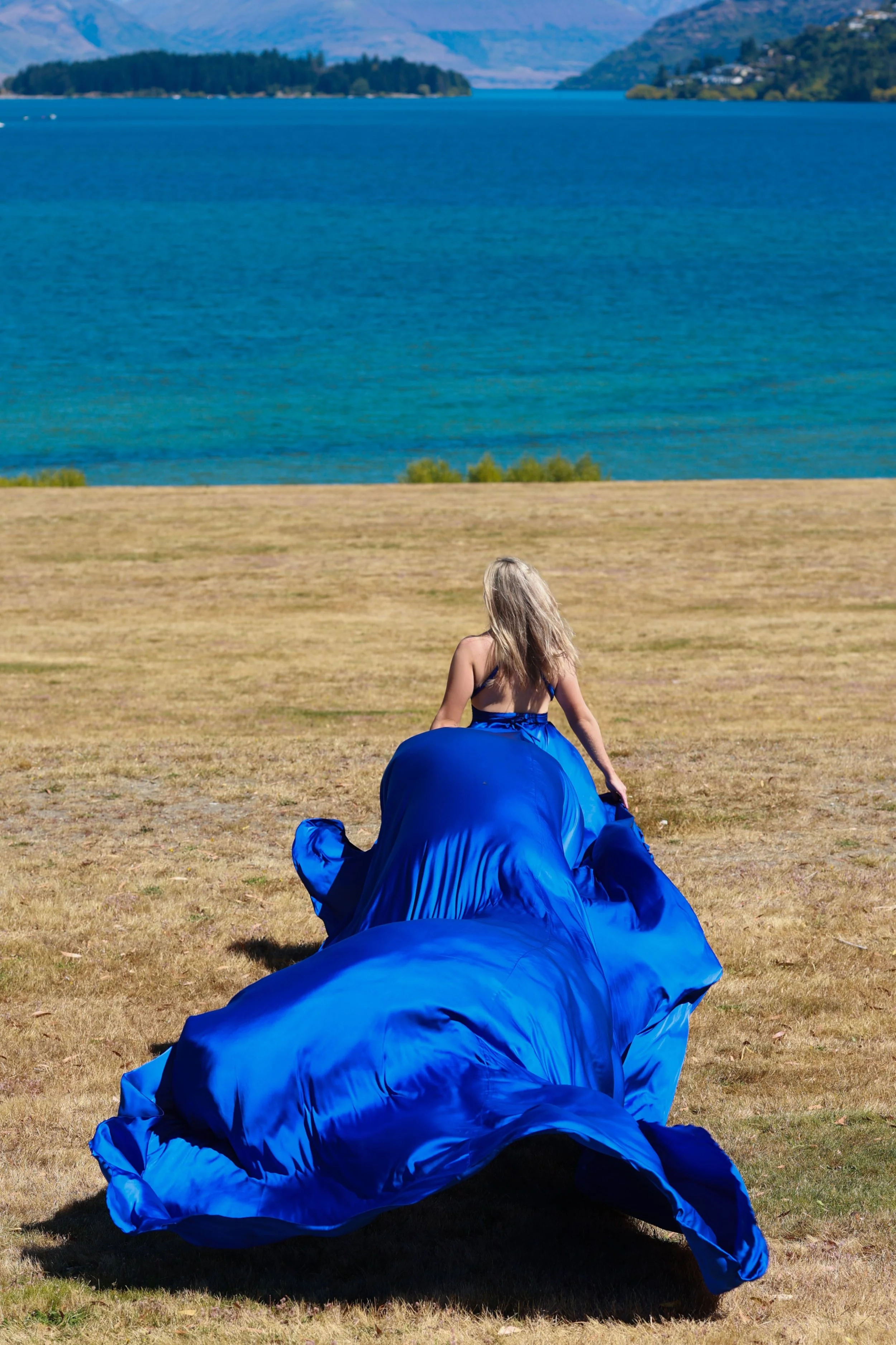 A woman in a blue dress and a blue paving bag sitting on dry grass near a large body of water with mountains in the background.