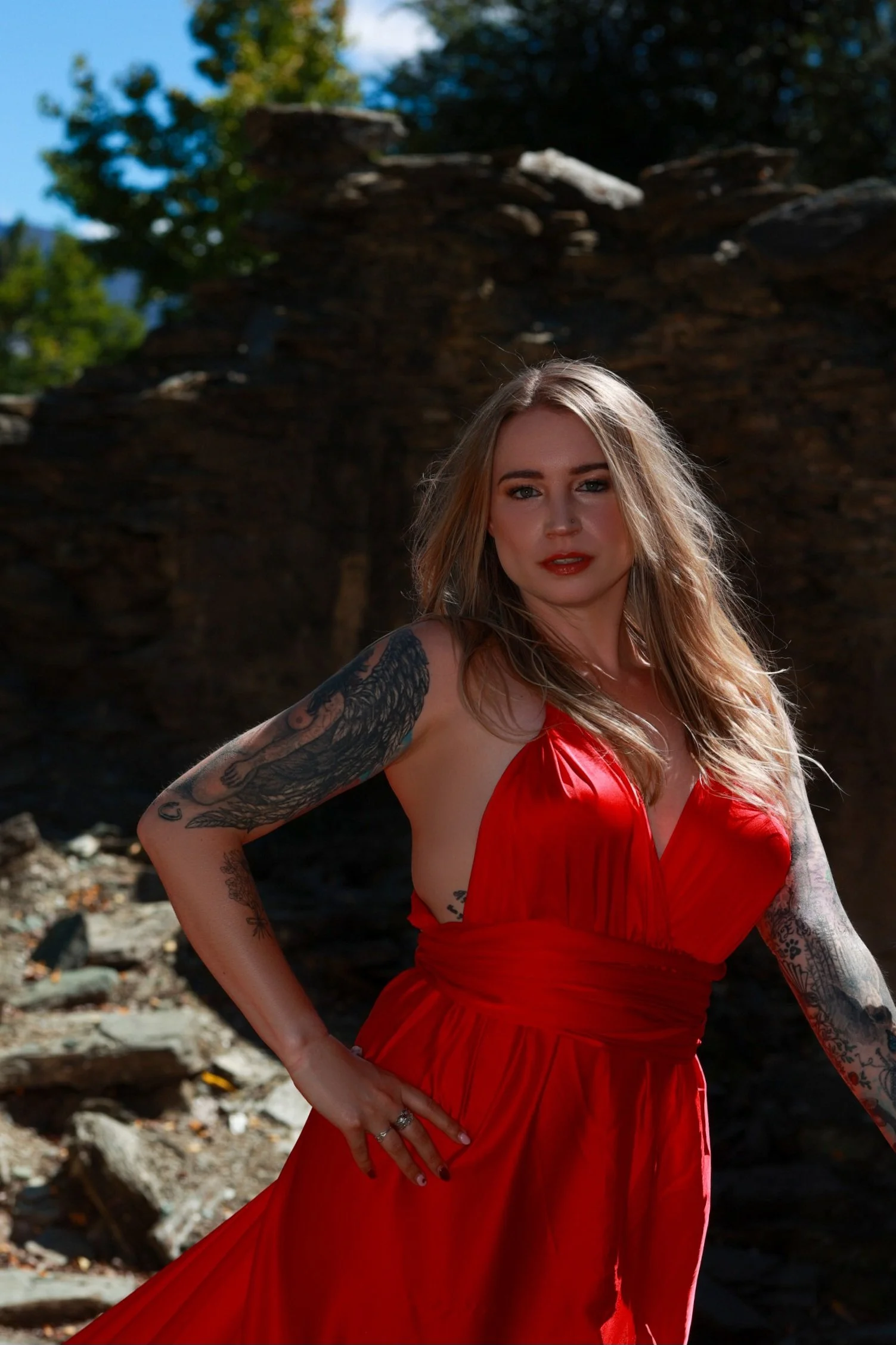 A woman in a red dress standing outdoors with a background of rocks and trees, with tattoos on her arms and a confident expression.