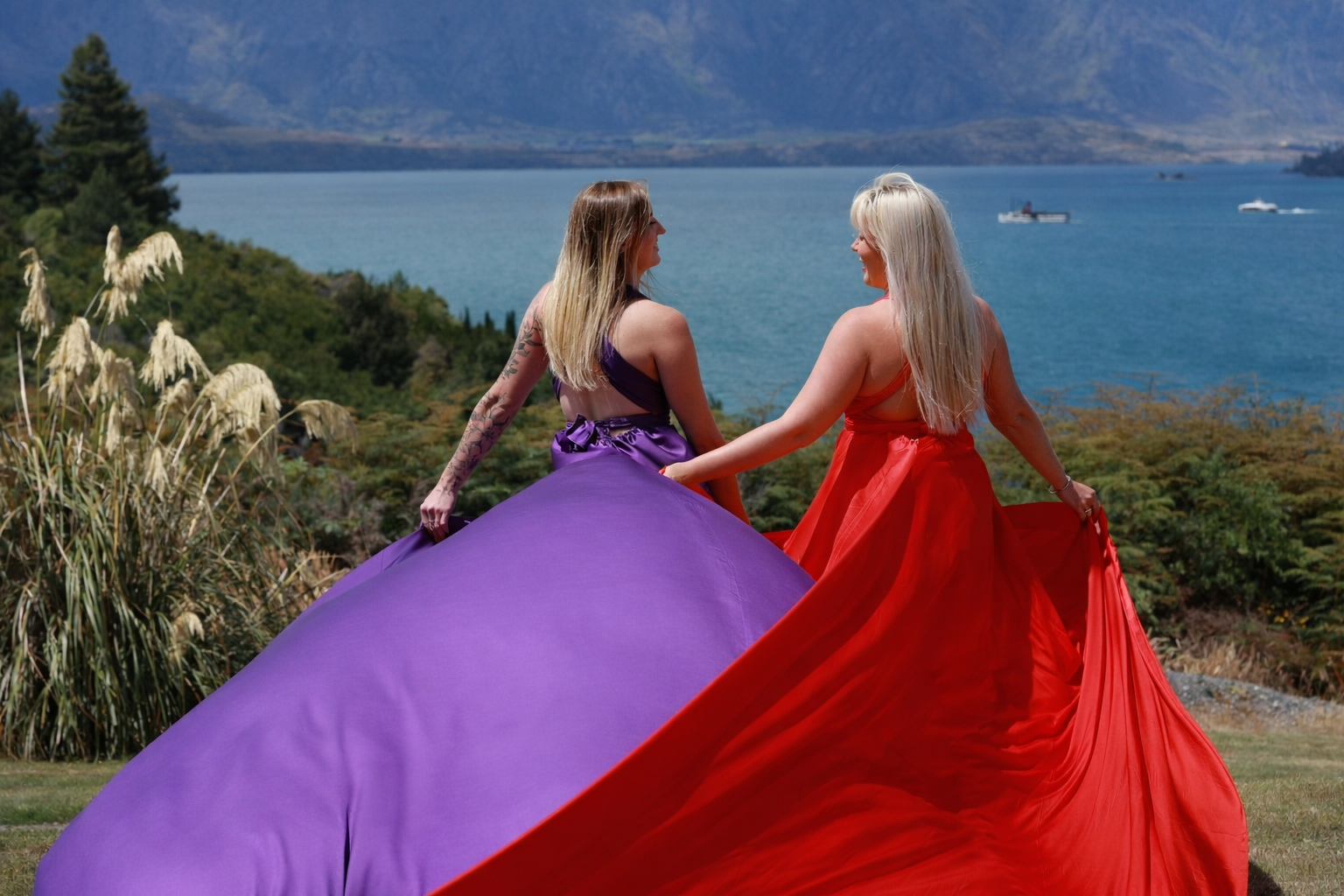 Two women in colorful evening gowns walking away from the camera near a lake with mountains in the background.