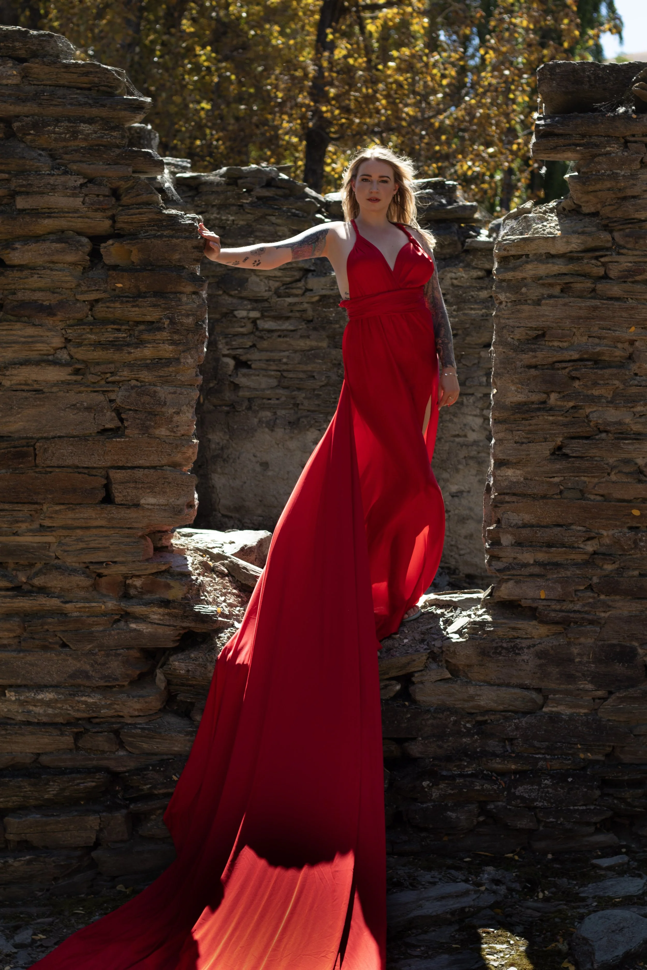 A woman in a long, flowing red dress standing among stone ruins with trees and autumn leaves in the background.