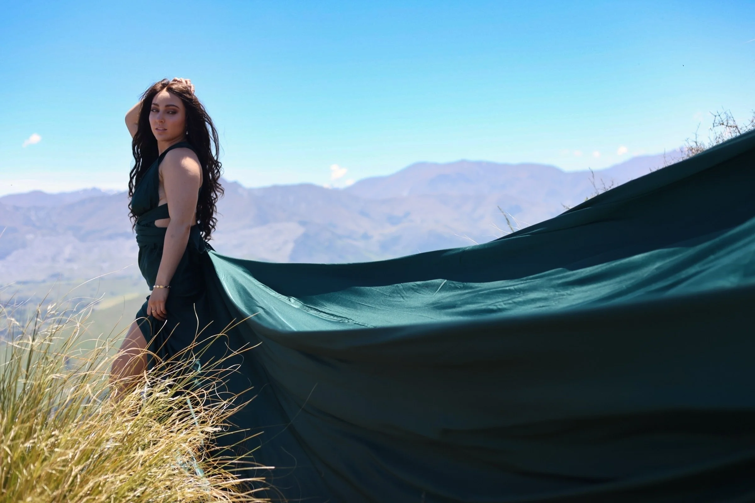 A woman with long dark hair wearing a black dress with cutouts stands outdoors on a grassy hill with mountains in the background, under a clear blue sky. She has one hand on her head and looks at the camera, with a flowing green fabric extending from