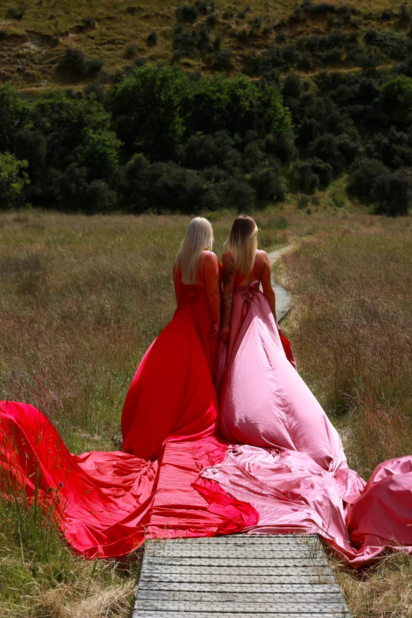 Two women in elegant dresses, one red and one pink, walking on a narrow path through a grassy field with hills and trees in the background.