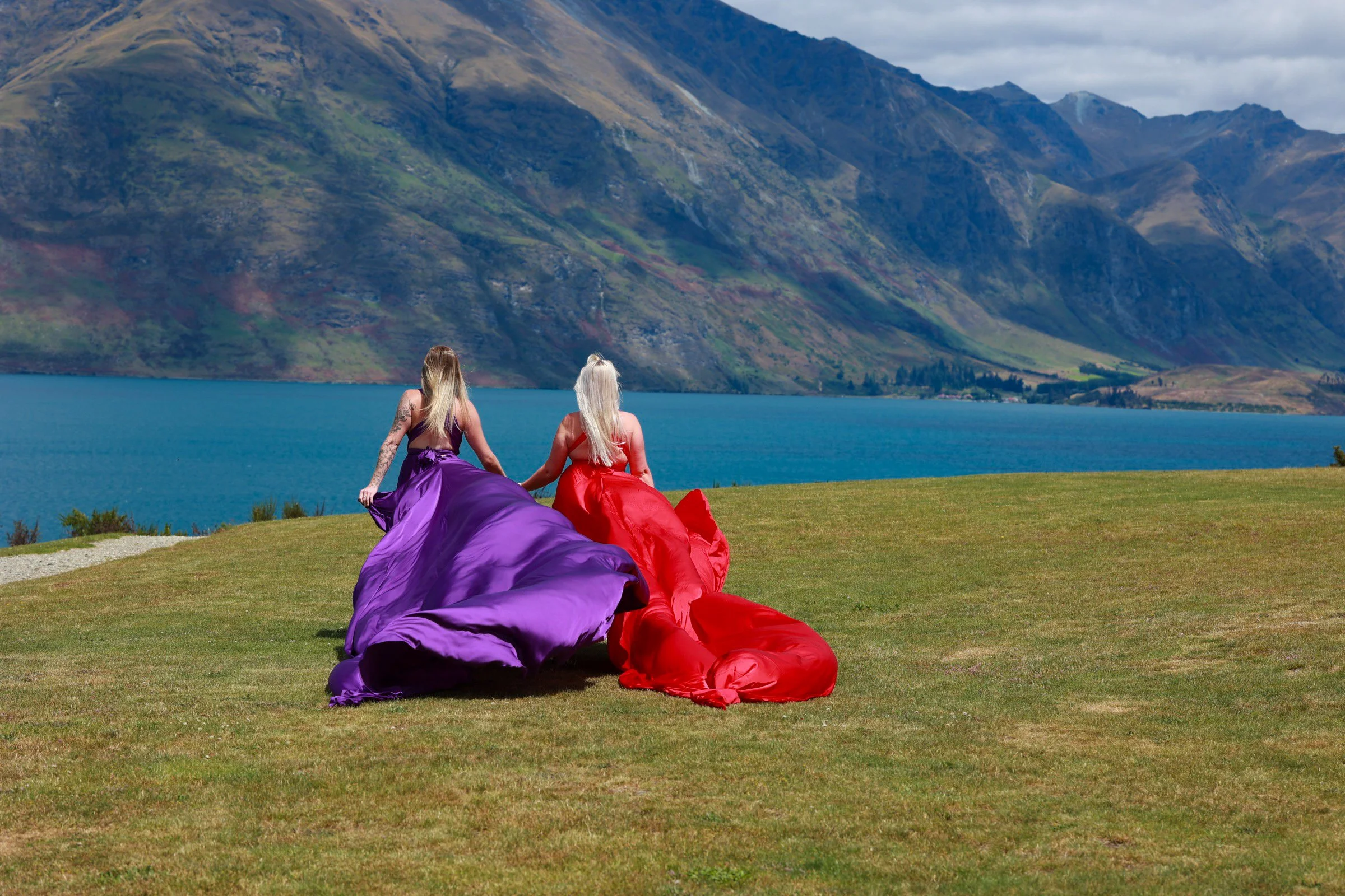Two women with long hair sitting on grass, wearing vibrant purple and red skirts, facing away, with a lake and mountains in the background.