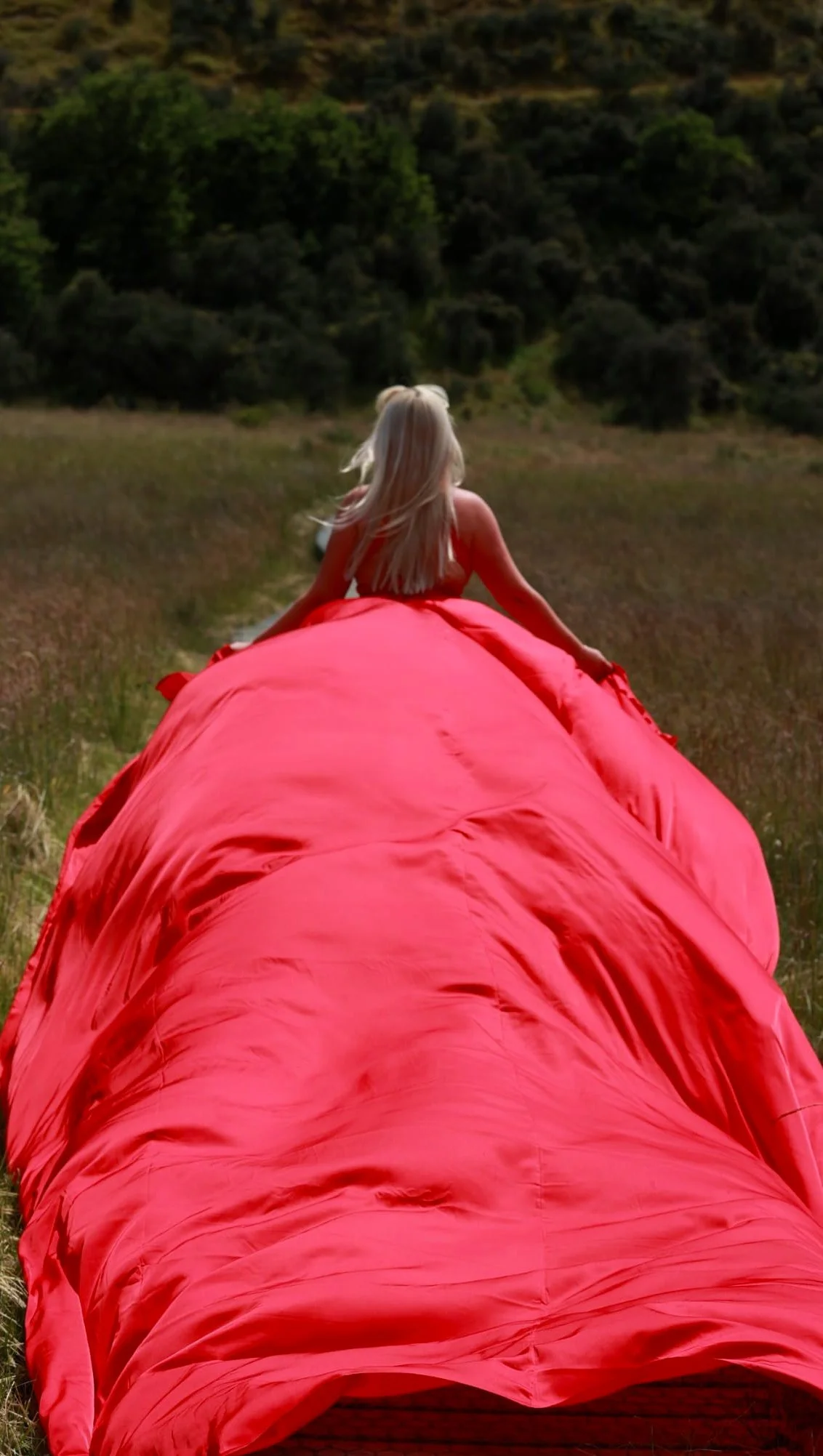 A person in a red jacket lying on a grassy field, looking toward a forested hillside in the distance.