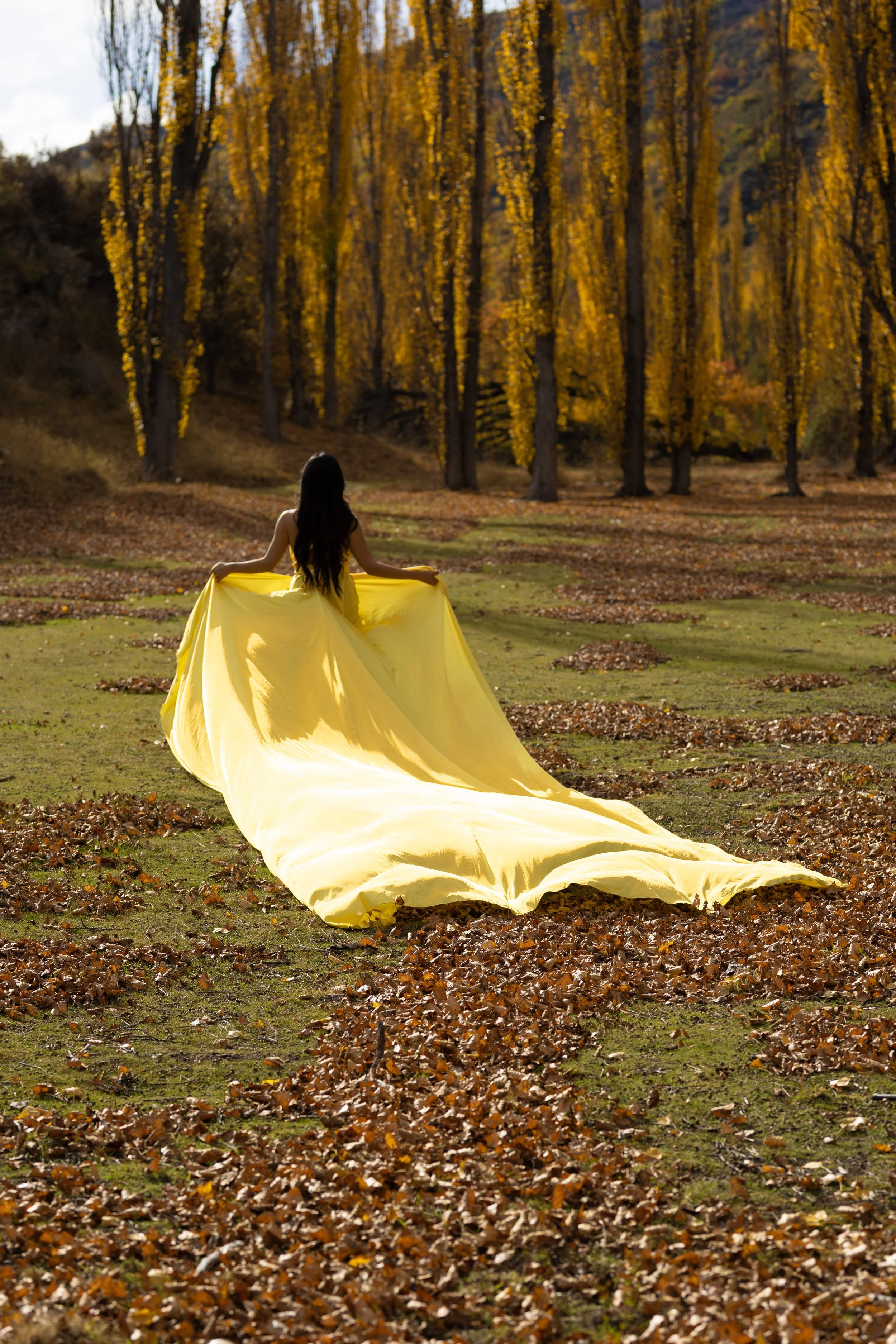 A woman in a flowing yellow dress standing in a park of autumn trees, with orange and yellow leaves on the ground and on the trees.