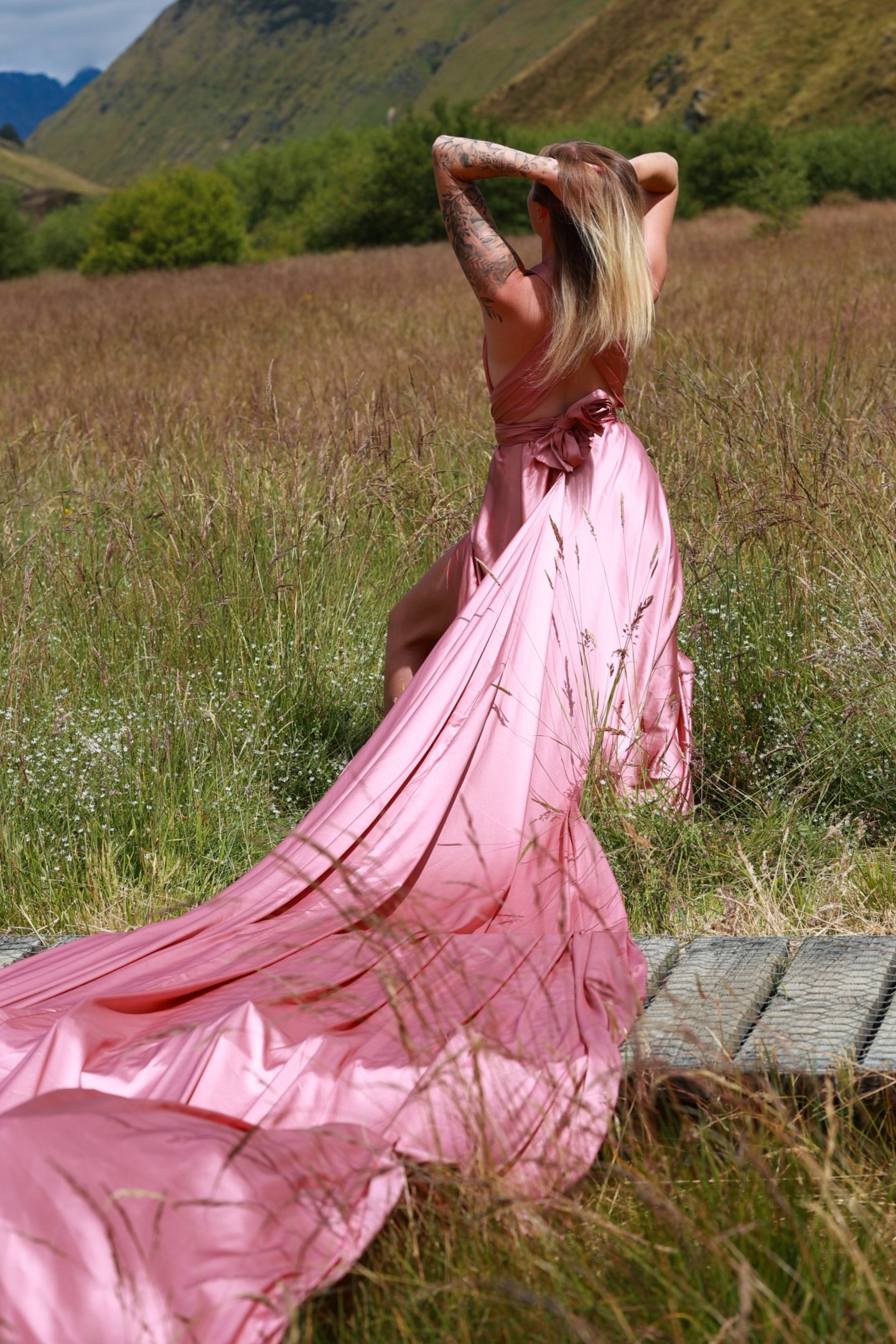 A woman wearing a pink dress with a train, standing in a field of tall grass with green hills and mountains in the background.