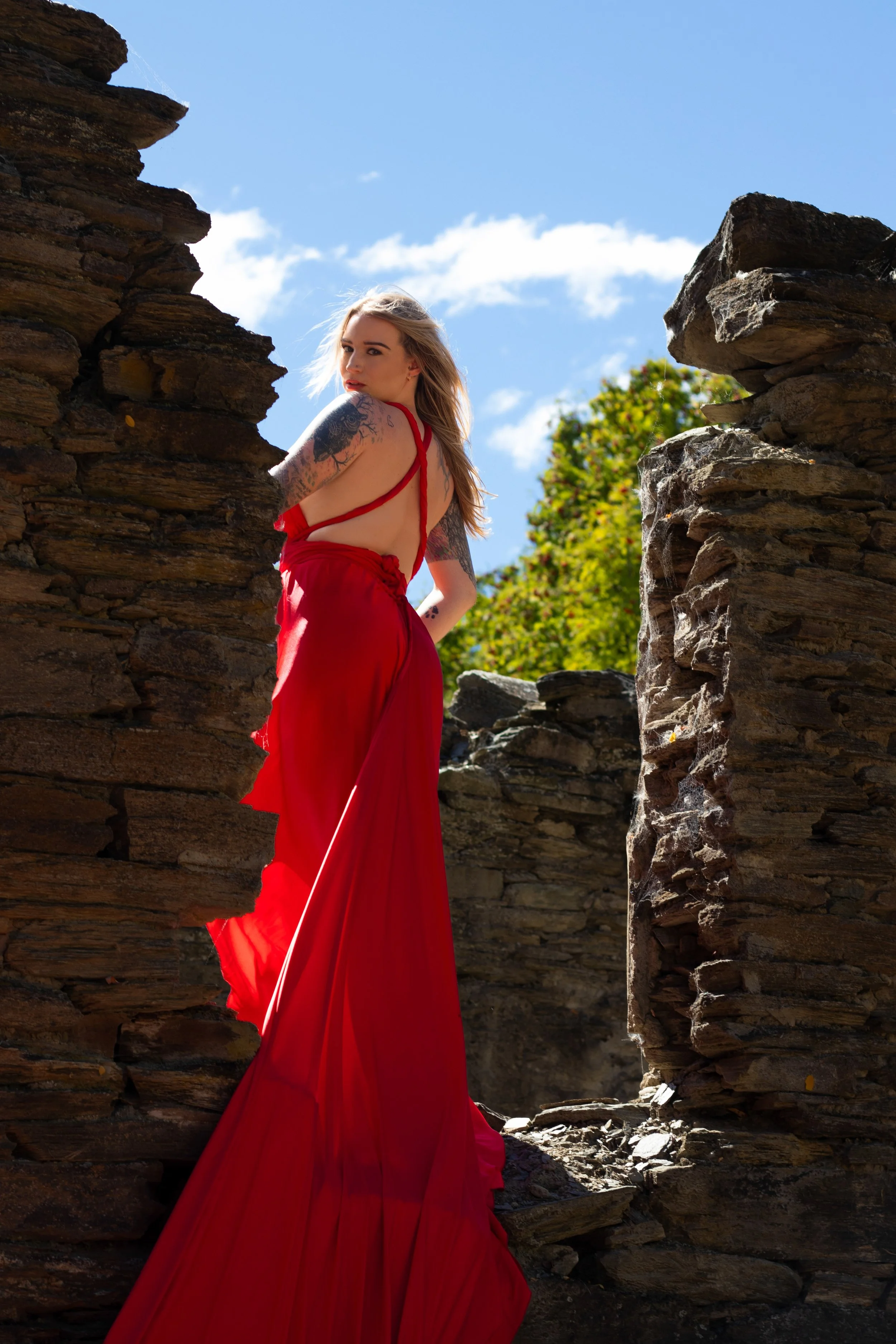 A young woman with long blonde hair stands between two large, weathered stone walls, wearing a flowing red dress with an open back. She looks over her shoulder toward the camera, with a tattooed arm visible. The sky is clear and blue, with green tree