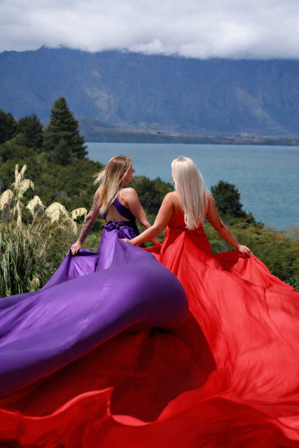 Two women in colorful dresses, one in purple and the other in red, holding their skirts and standing outdoors near a lake with mountains and cloudy sky in the background.