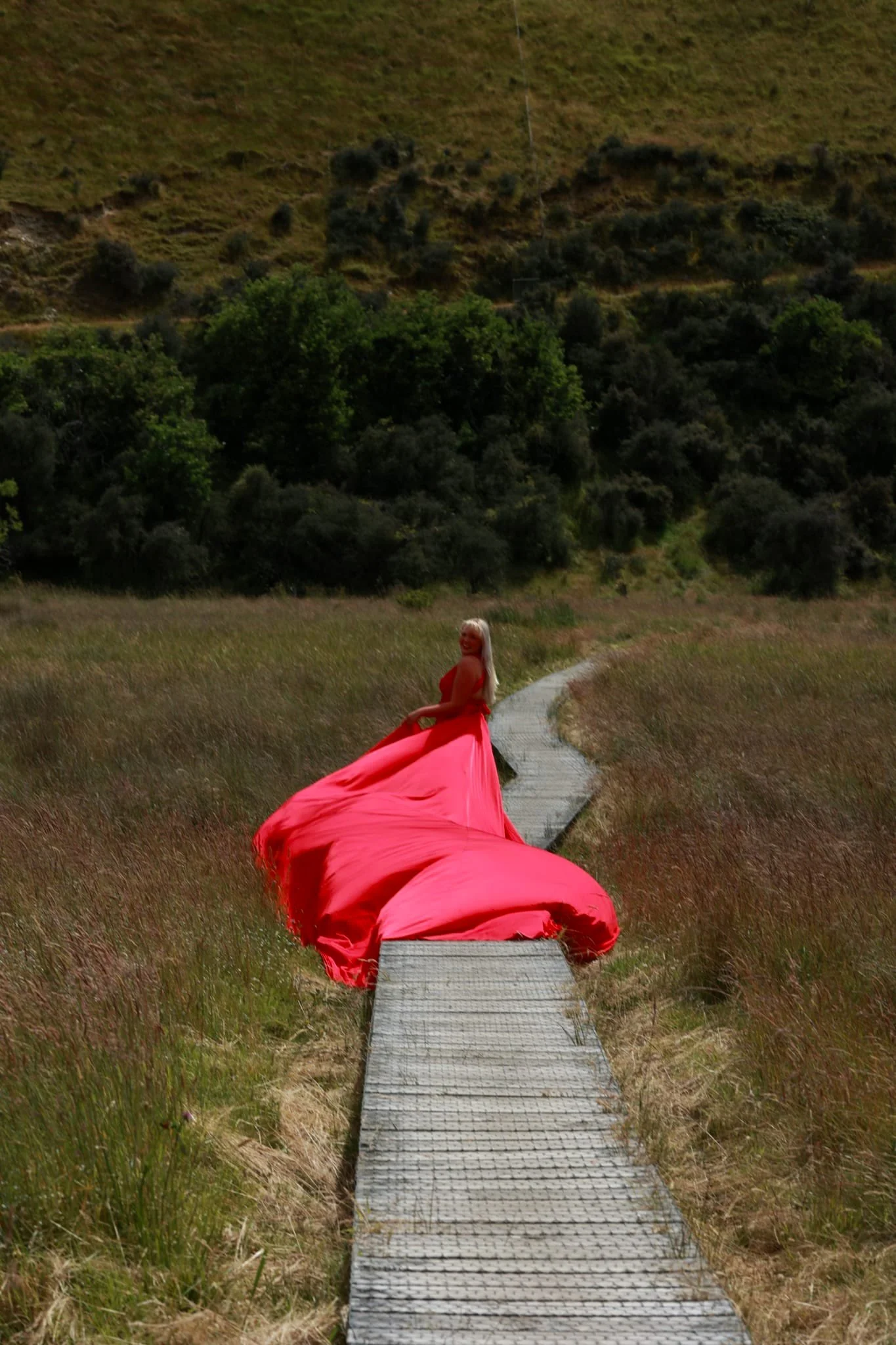 A woman in a pink dress sitting on a wooden pathway in an open grassy field with trees and hills in the background.