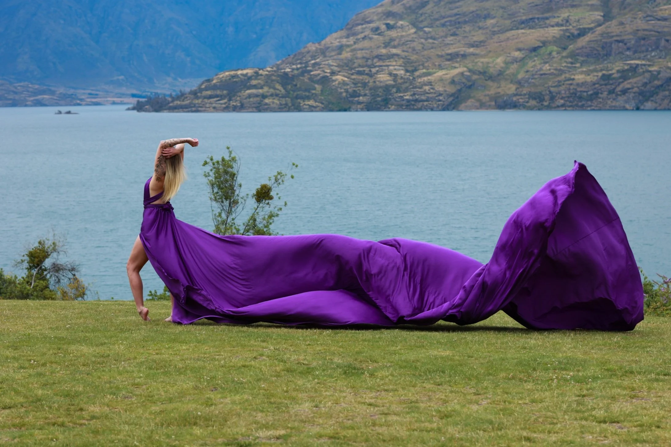 A woman with long blonde hair and tattoos on her left arm, dressed in a purple dress, is standing outdoors on grass near a body of water with mountains in the background. She holds her hair back with her left hand while standing on her right leg. A l