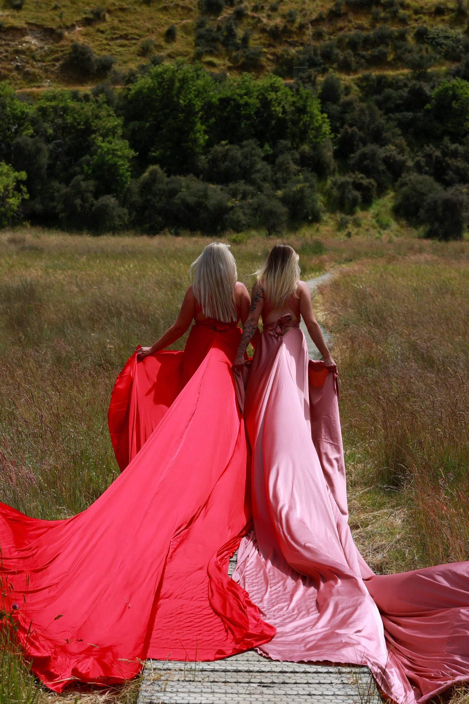 Two women in colorful gowns, one in red and the other in pink, walking away from the camera on a grassy path in a field with trees and hills in the background.