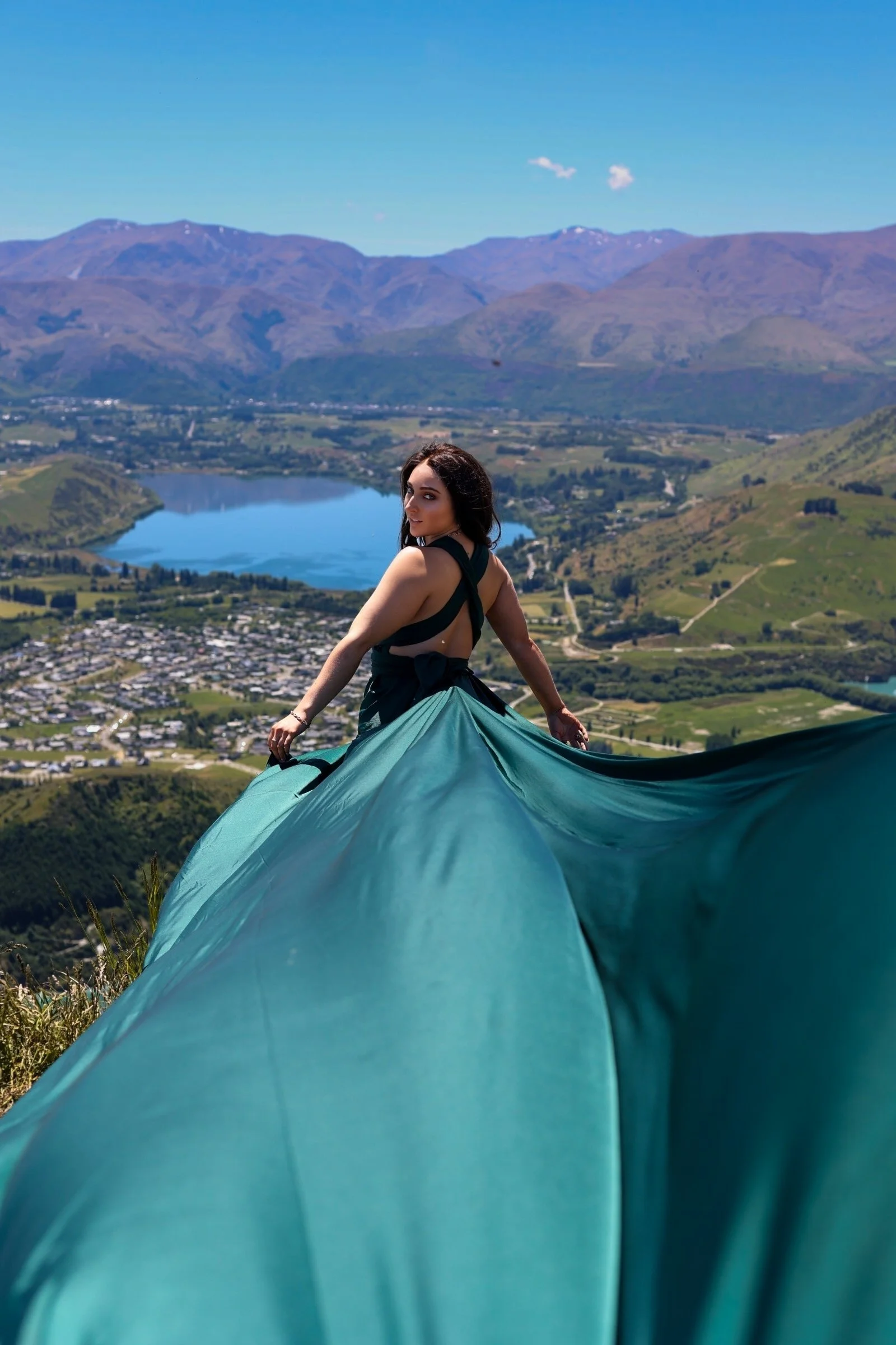 A woman in a black top and teal skirt standing on a hill with a panoramic view of a landscape including a lake, green fields, and mountains under a clear blue sky.