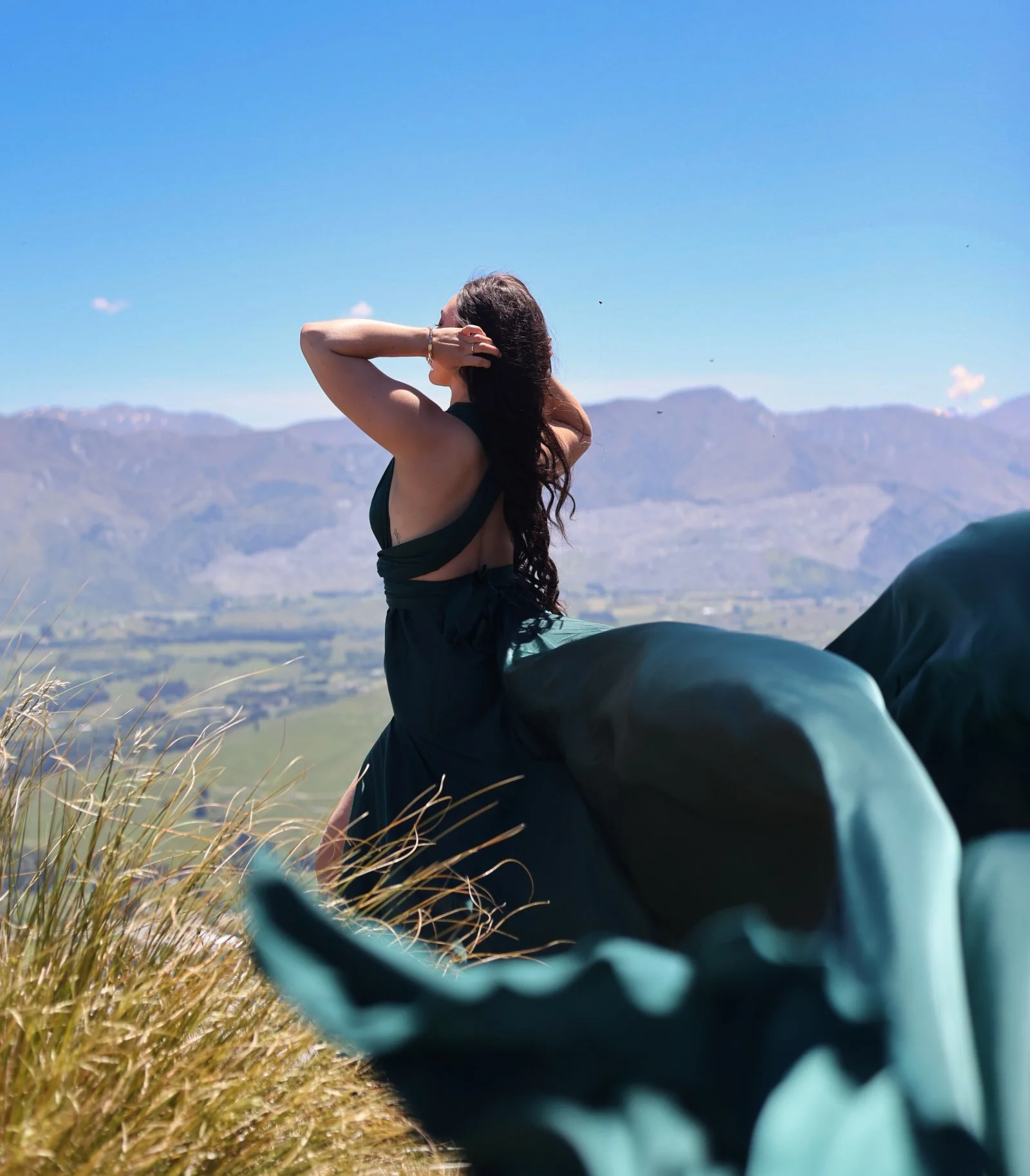 A woman with long dark hair is sitting outdoors on grassy terrain, facing away from the camera, with mountains and a blue sky in the background. She has one arm raised and bent behind her head, and the other arm across her waist. She is wearing a sle